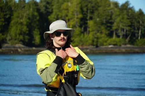 Person in a yellow jacket and hat adjusting gear on a boat with water and forest in the background