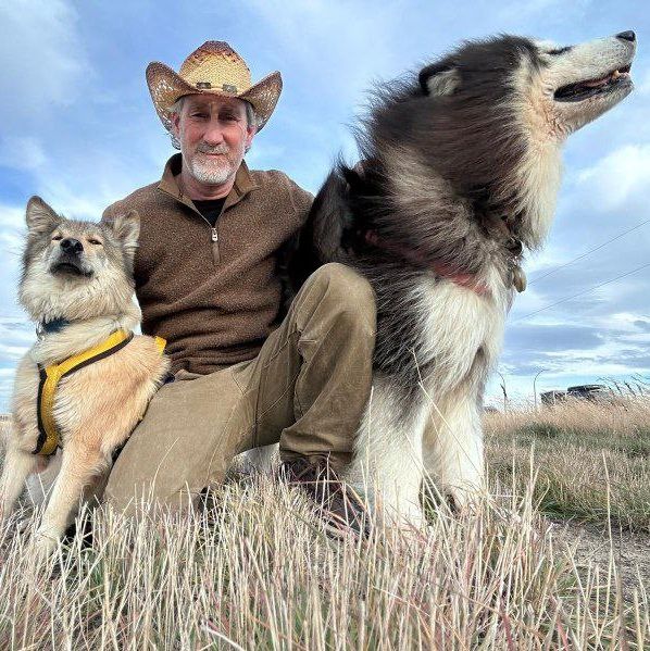 Man in a cowboy hat sitting in tall grass with two large dogs under a cloudy sky