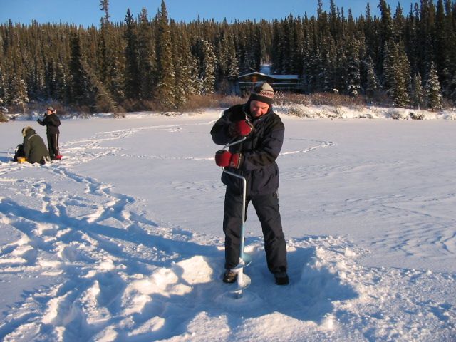 Person in dark winter gear holding a ski pole on a snowy frozen lake, with pine trees in the background