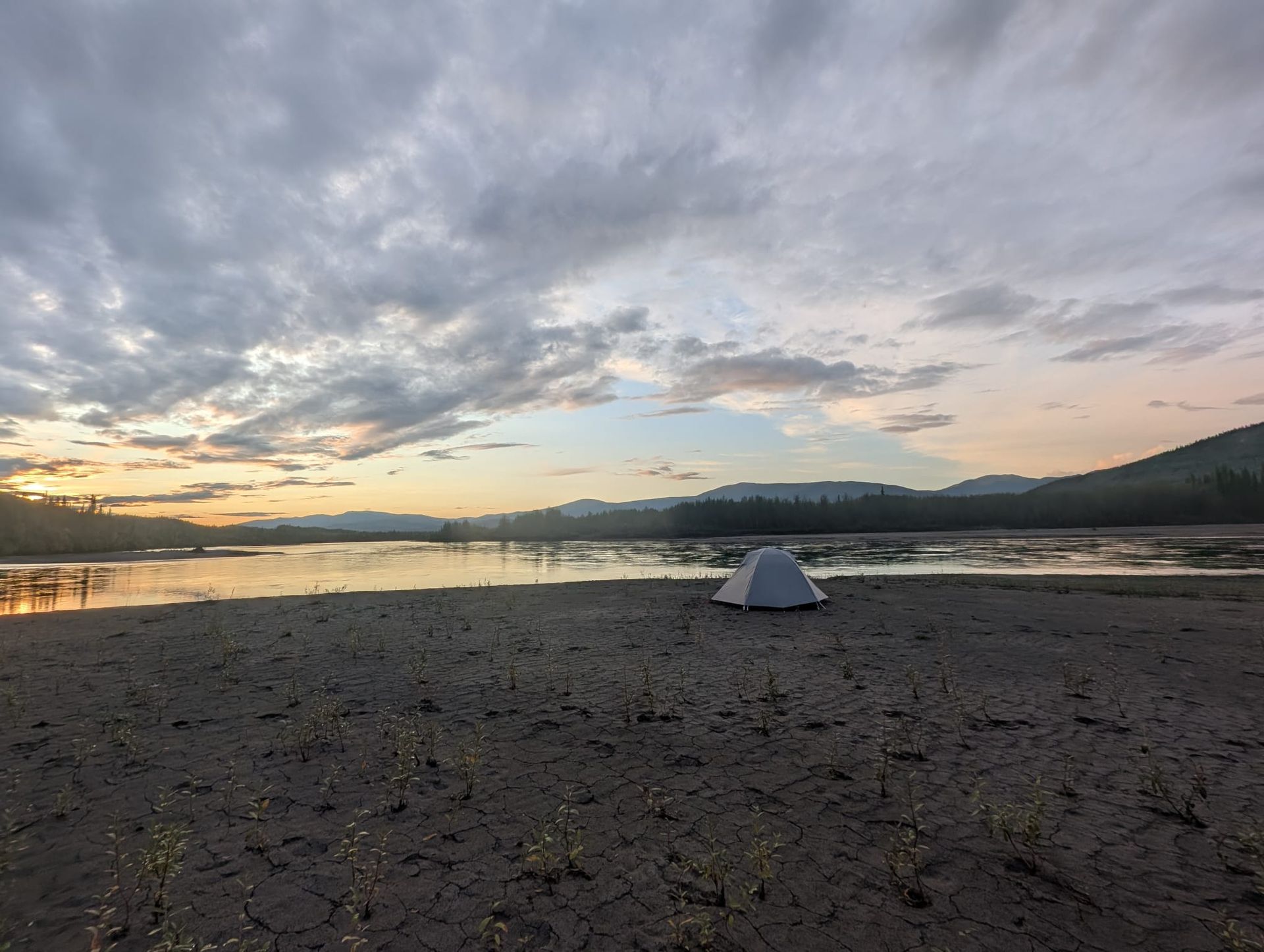 Tent on a rocky lakeshore at dusk, with calm water, distant hills, and a cloudy sky.