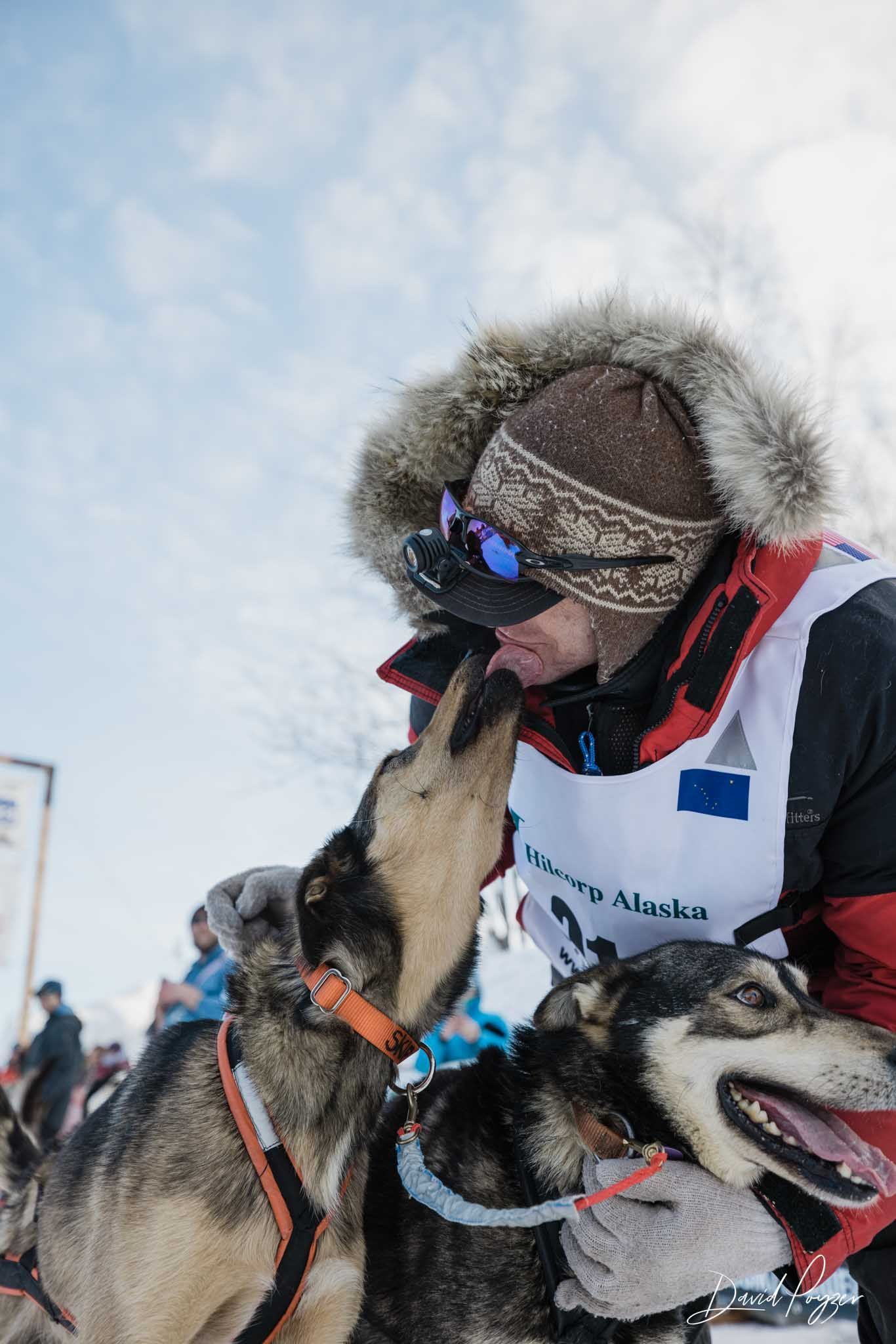 Musher in fur hood hugs two huskies in snowy Arctic conditions