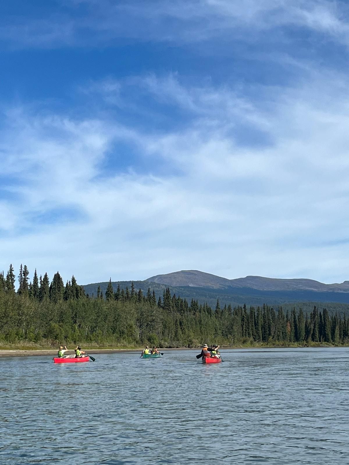 Three kayaks on a calm lake with forested shore and mountains under a blue sky