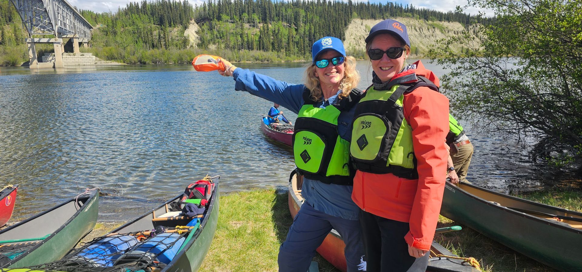 Two people in life jackets posing beside kayaks on a lakeshore, one pointing across the water.