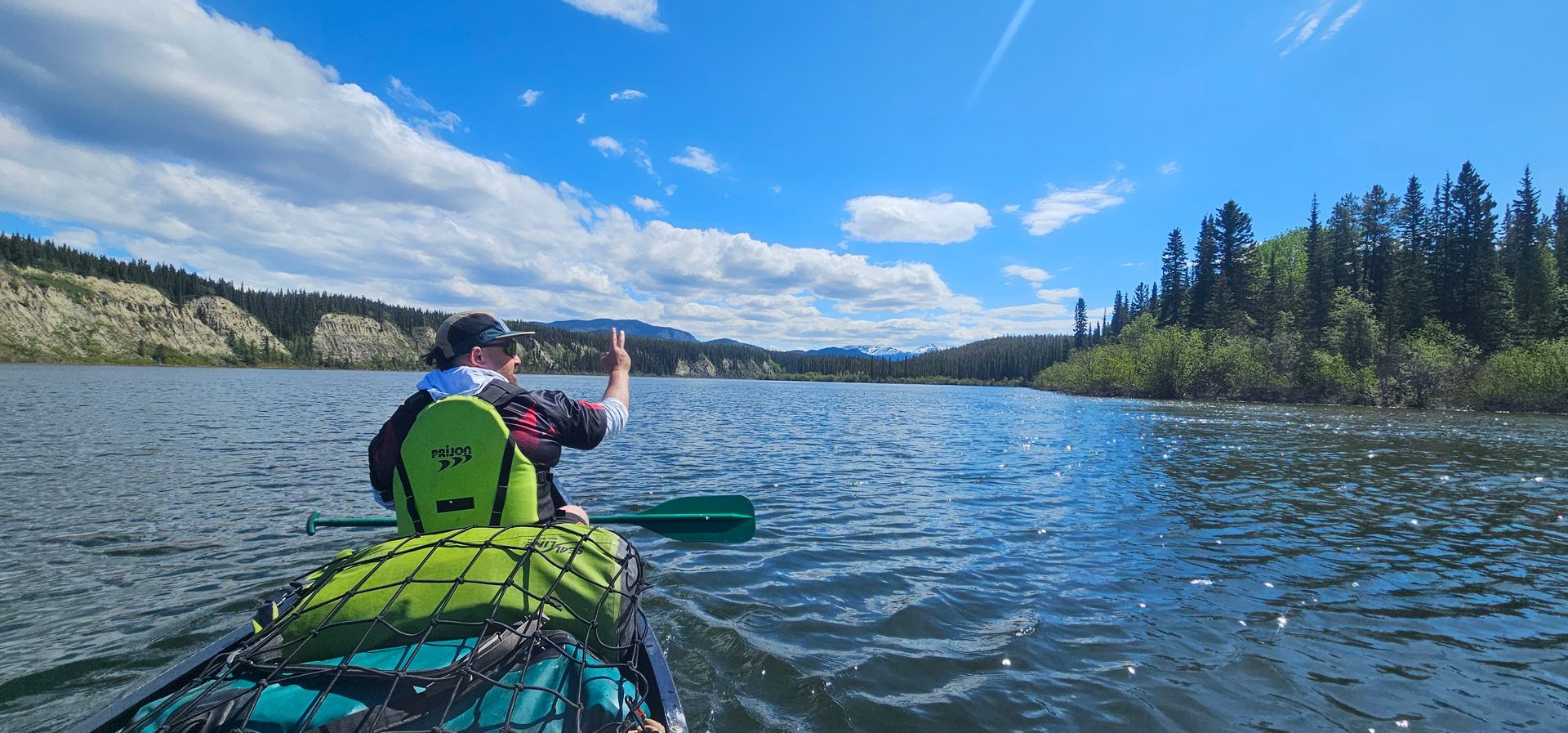 Kayaker in a green vest paddles a blue kayak on a lake surrounded by forested hills under a cloudy sky