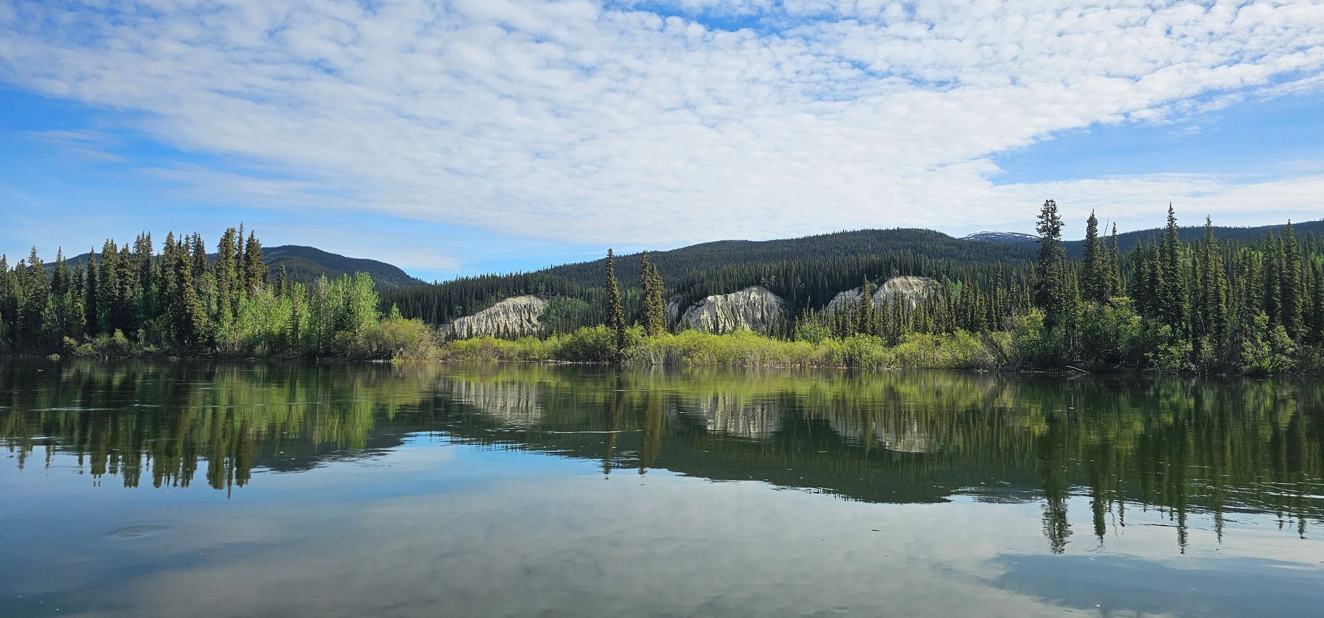Calm lake reflecting forested hills and blue sky under scattered clouds