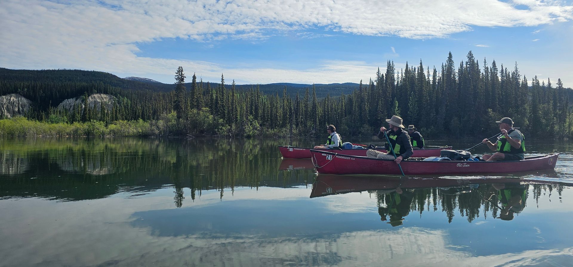 Red canoe with people on a calm lake, reflected mountains and forest in the background.