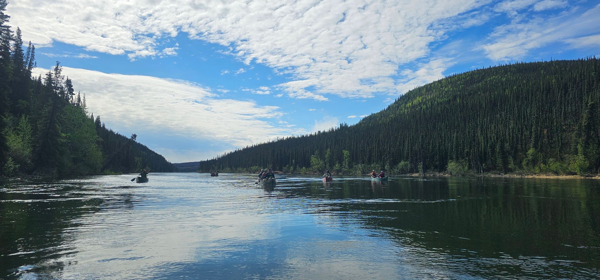 Calm forested lake under a cloudy blue sky, with distant boats and reflections on the water
