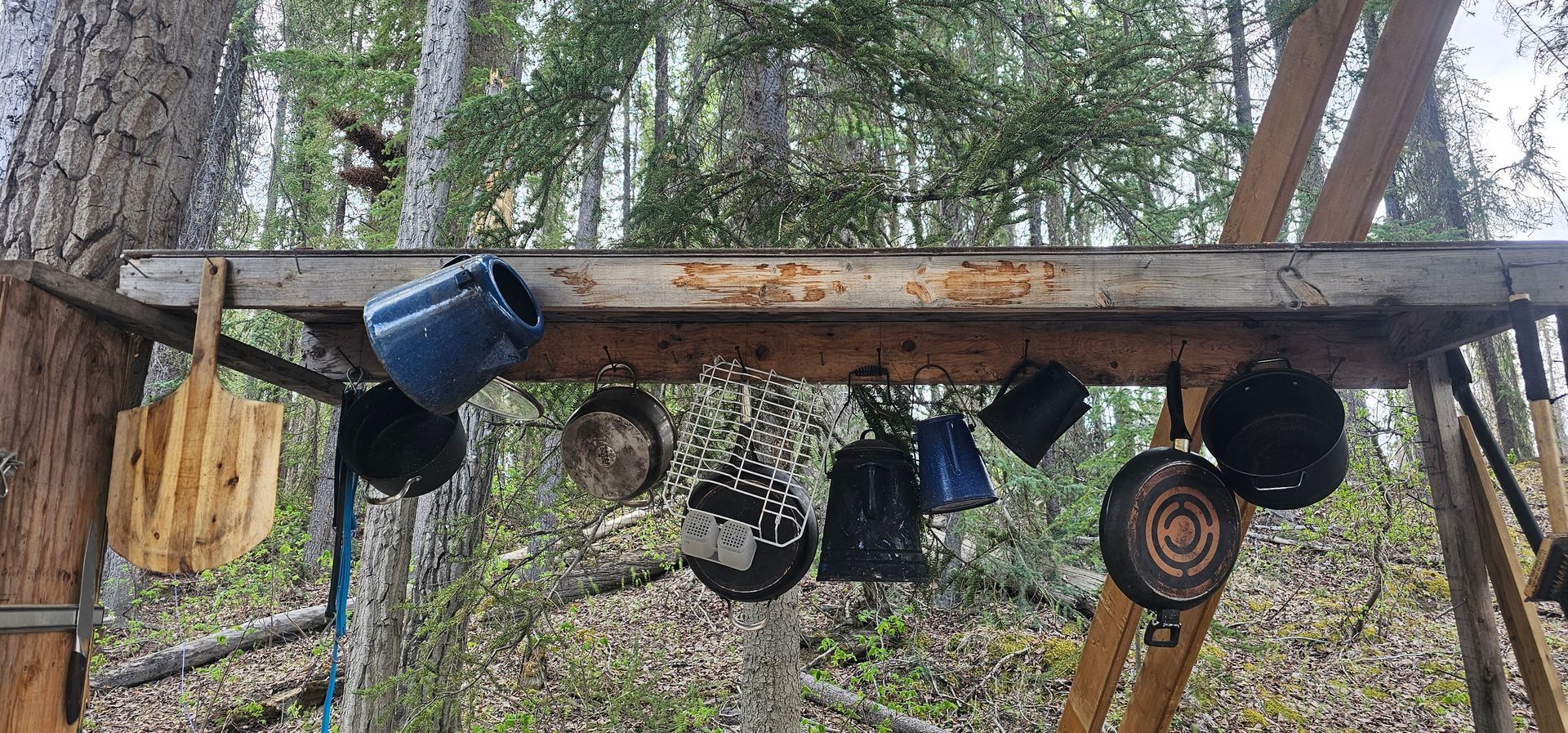 Rustic outdoor shelf with hanging mugs, pots, and a circular target against a wooded backdrop