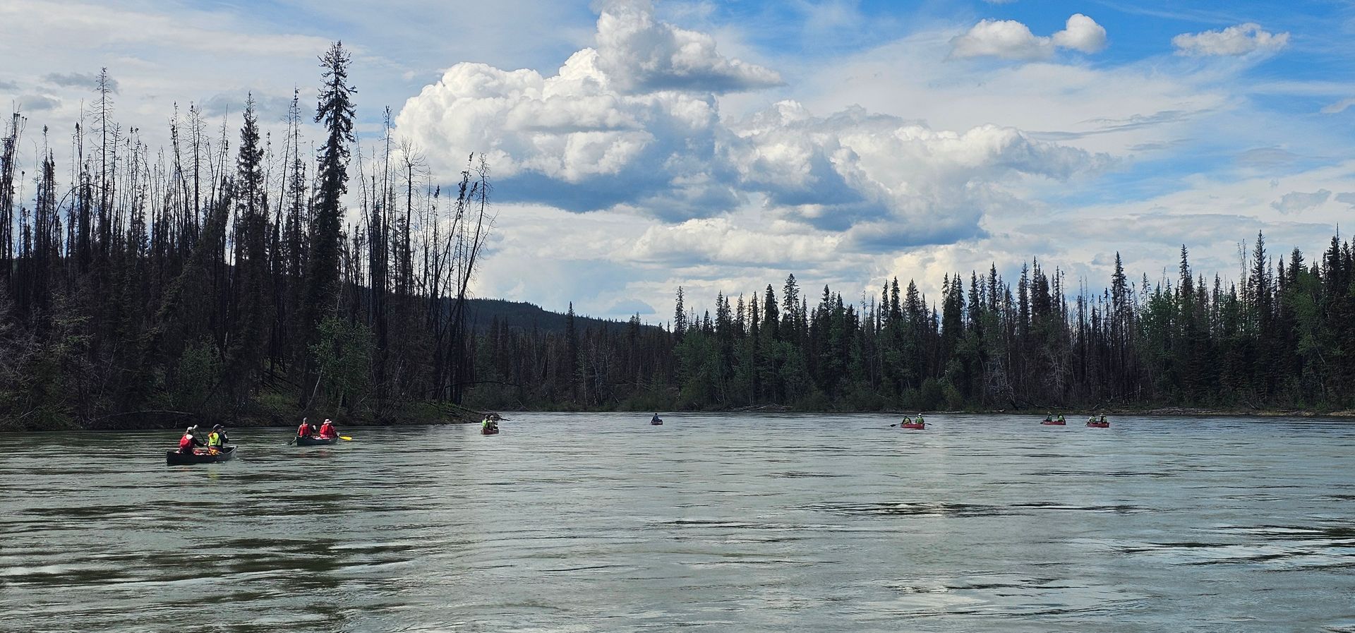 Calm river with two kayakers near a forested shoreline under cloudy skies