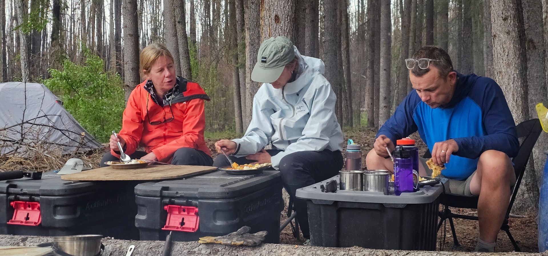 Three people in outdoor gear cooking at a campsite by a fire pit in a wooded area