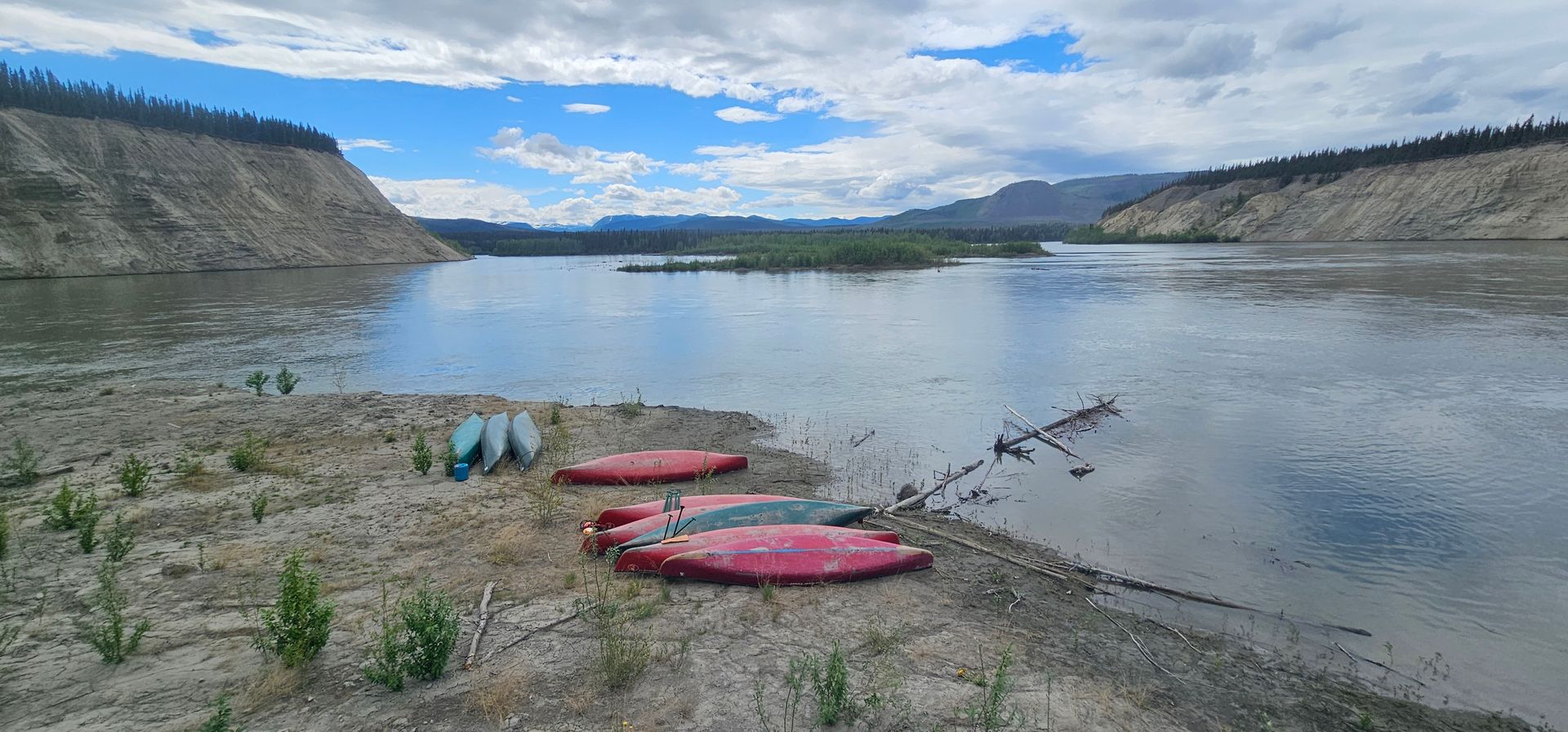Kayaks on a sandy riverbank beside calm water, with cliffs and cloudy sky in the background