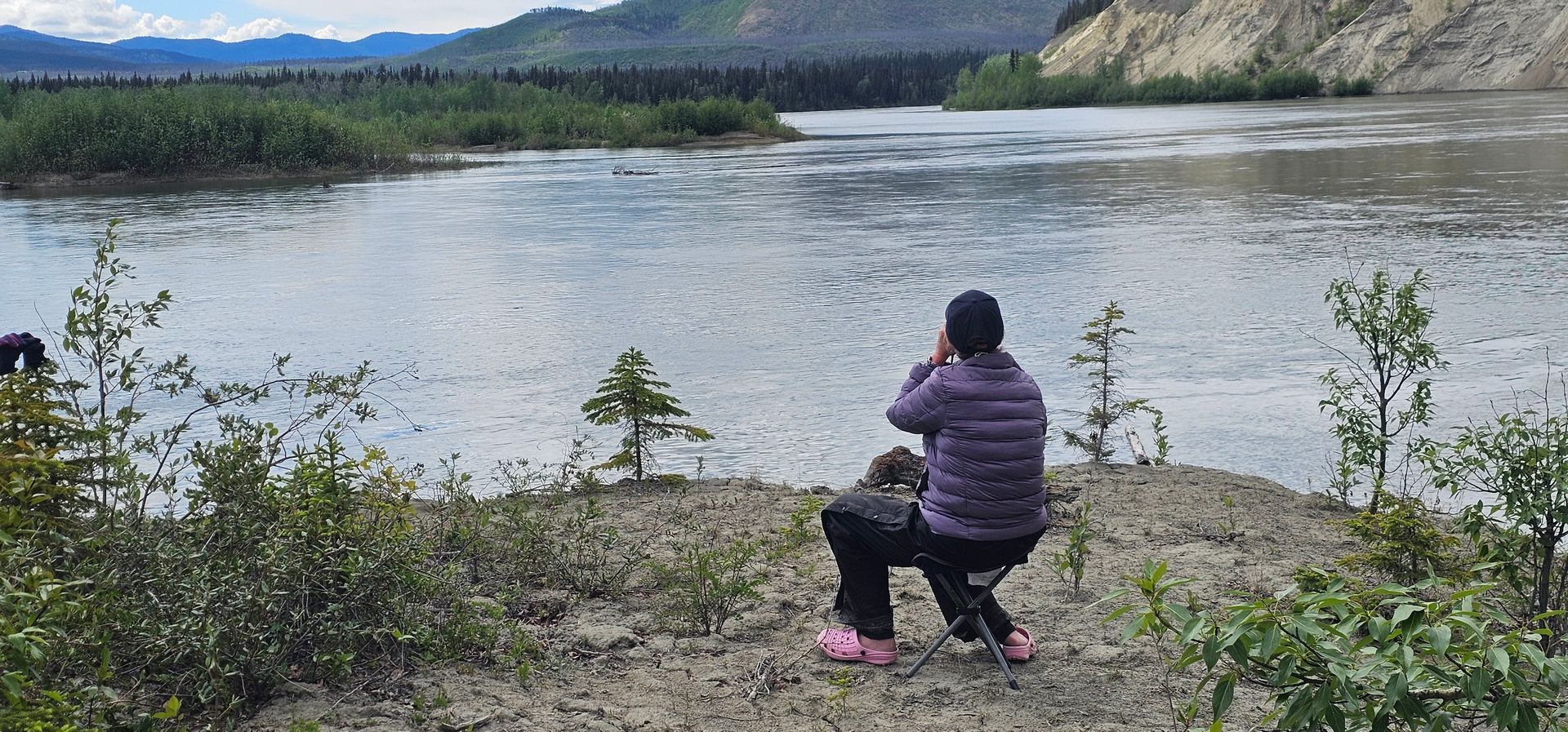 Person in a purple jacket photographing a river and mountains from a rocky shore