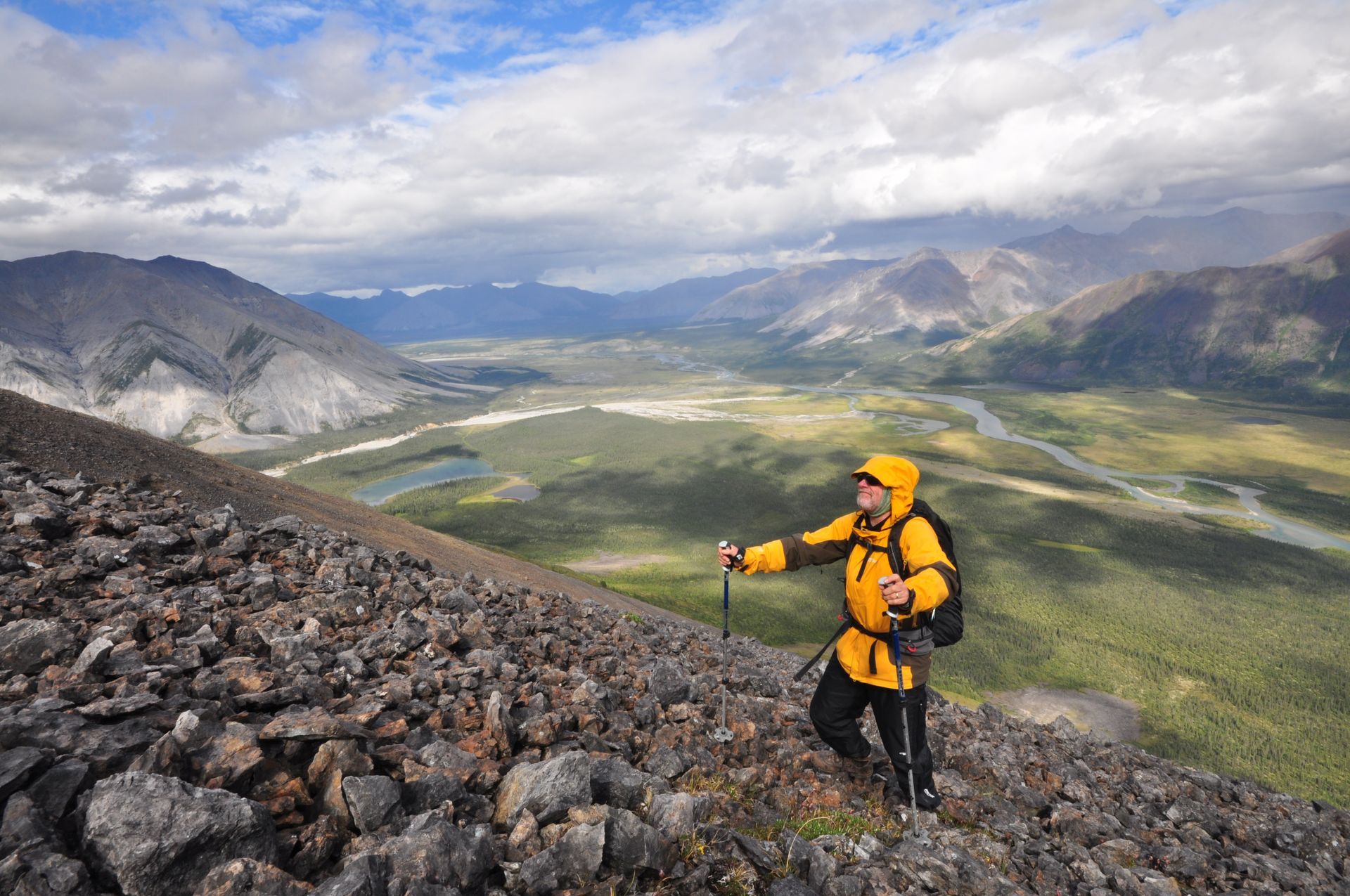 Hiker in yellow gear on rocky mountain slope overlooking a wide green valley and distant peaks