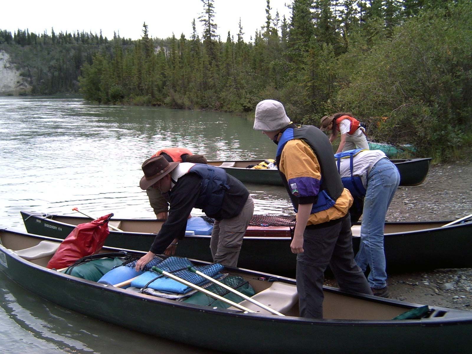 People loading canoes by a river, with gear and backpacks on board.