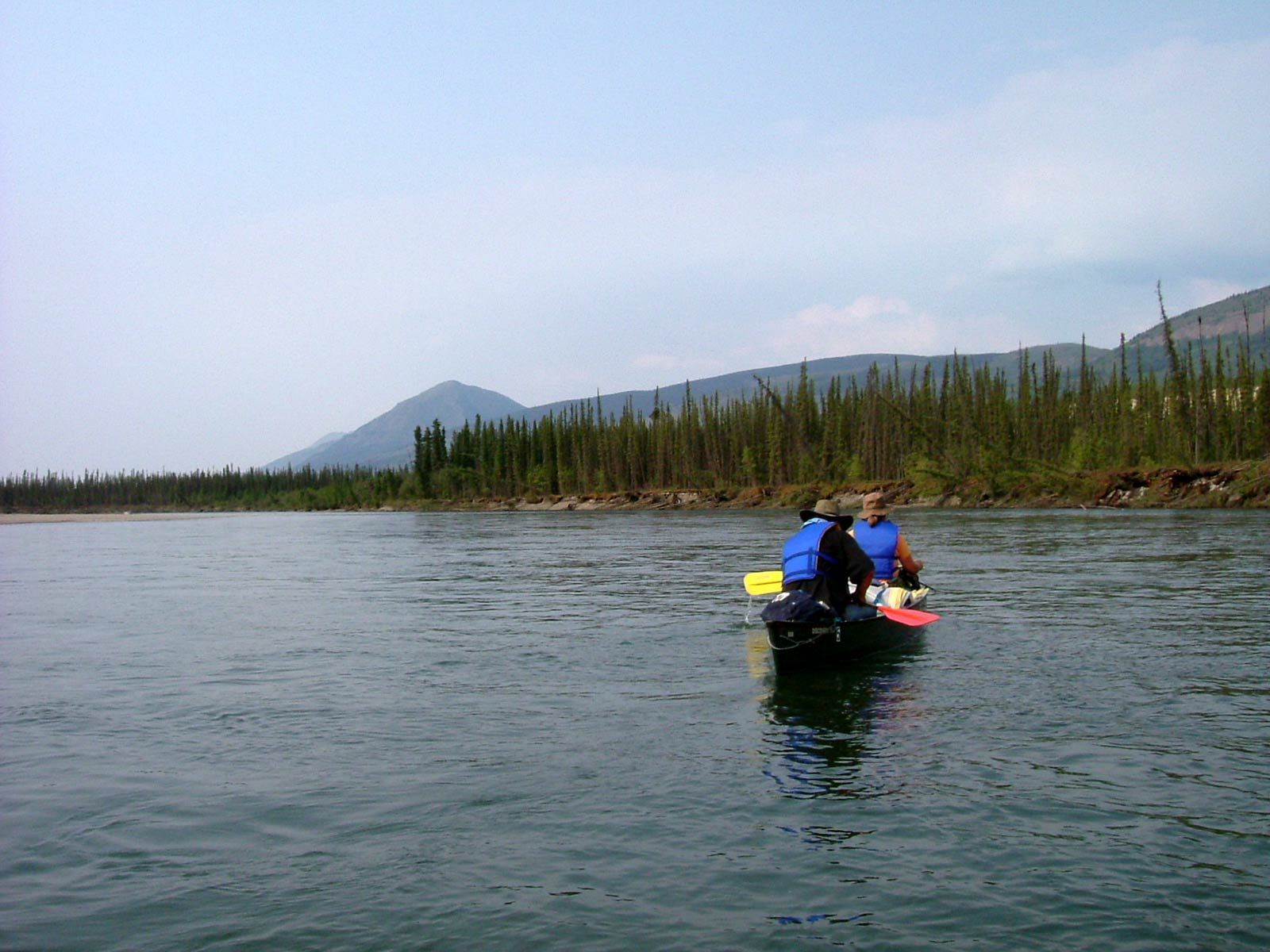 Two people in a small kayak on a calm river with mountains and trees in the distance.