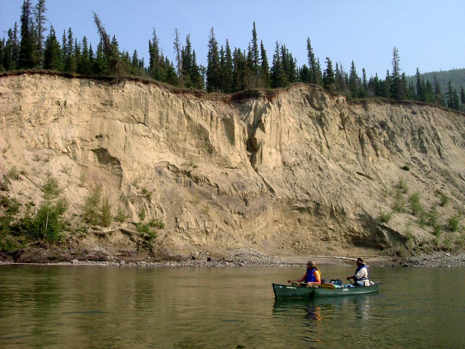 Two people in a green canoe on a calm river below a steep sandy cliff with pine trees.