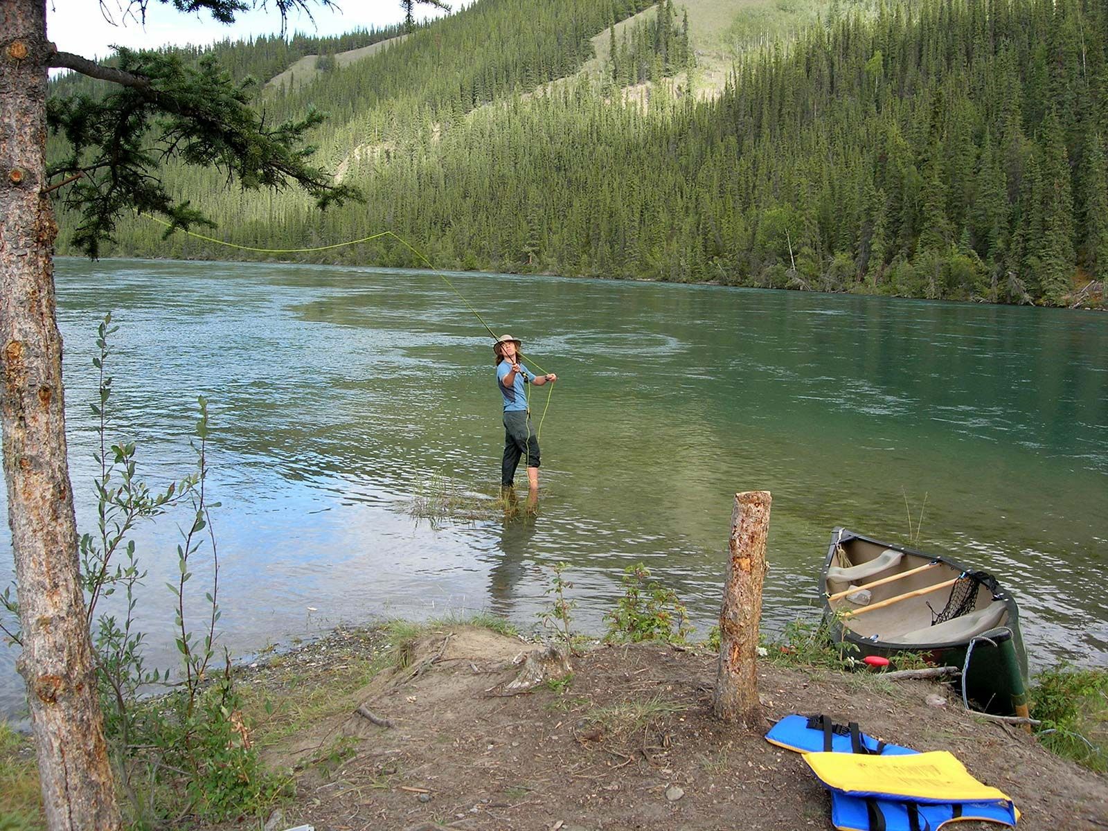 Person standing in a river beside a canoe, with forested hills in the background.