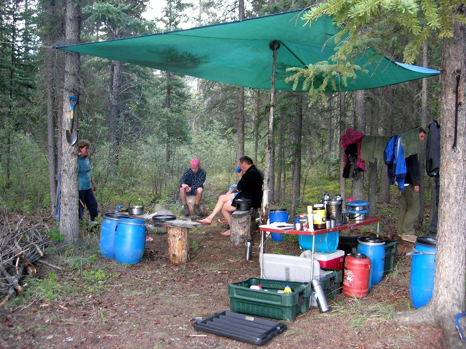People camping under a green tarp in a forest, with cooking gear, barrels, and hanging clothes.