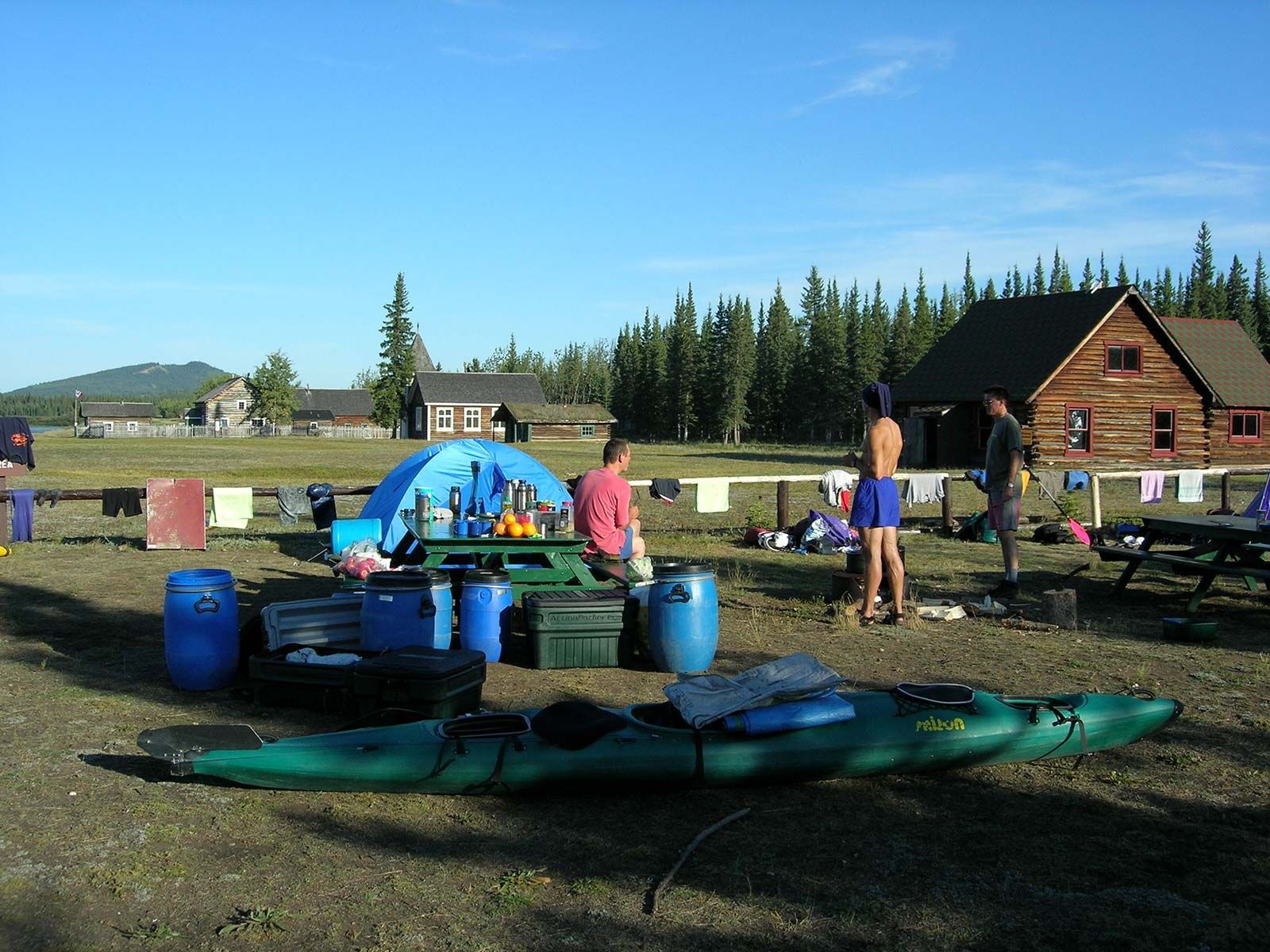 People on a lakeside picnic area with kayaks, barrels, and cabins in the background.