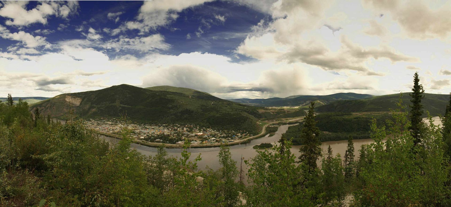 Mountain valley with river, green hills, and cloudy sky under a bright landscape.