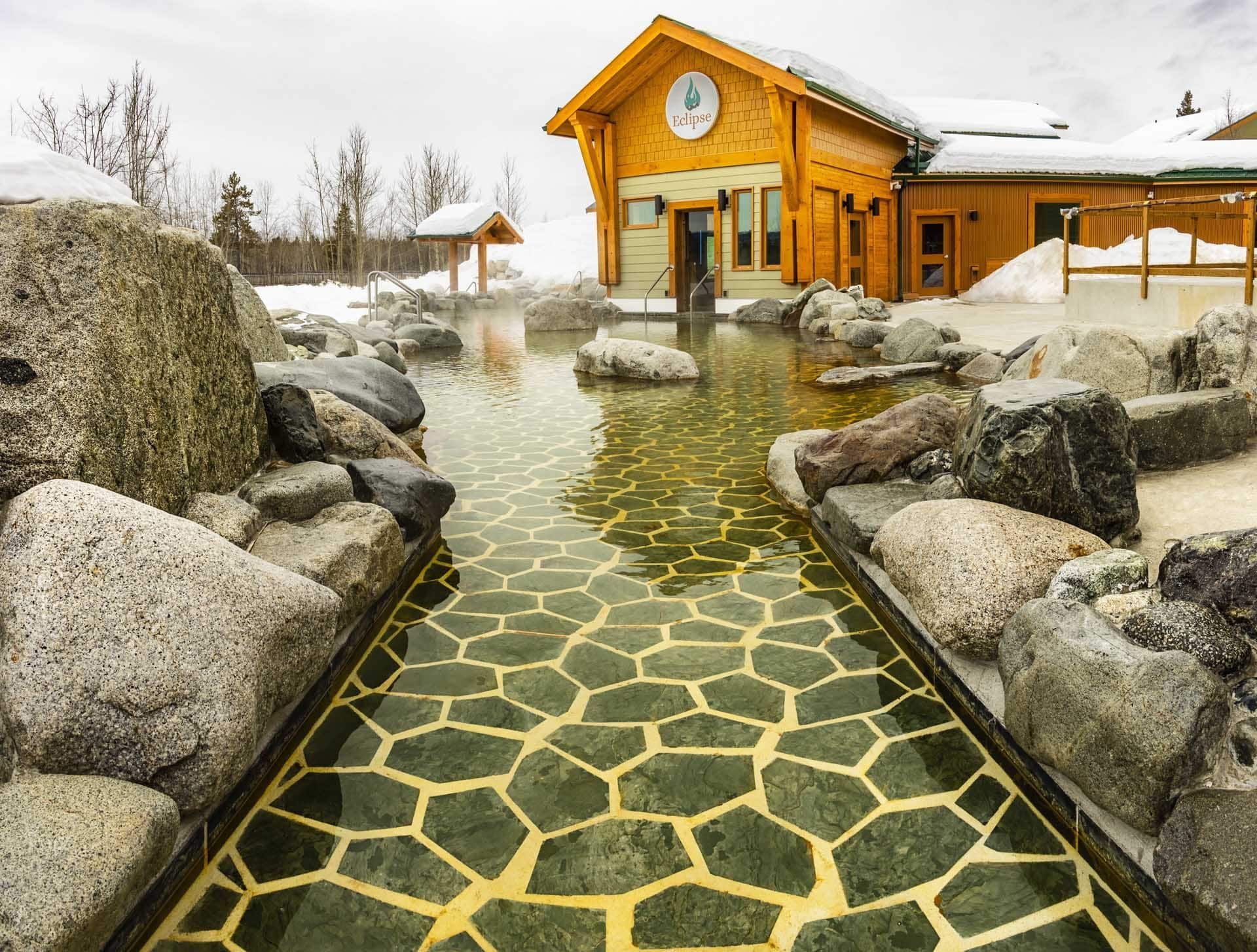 Outdoor hot spring path with hexagonal stone tiles leading to a wooden building amid large rocks and steam