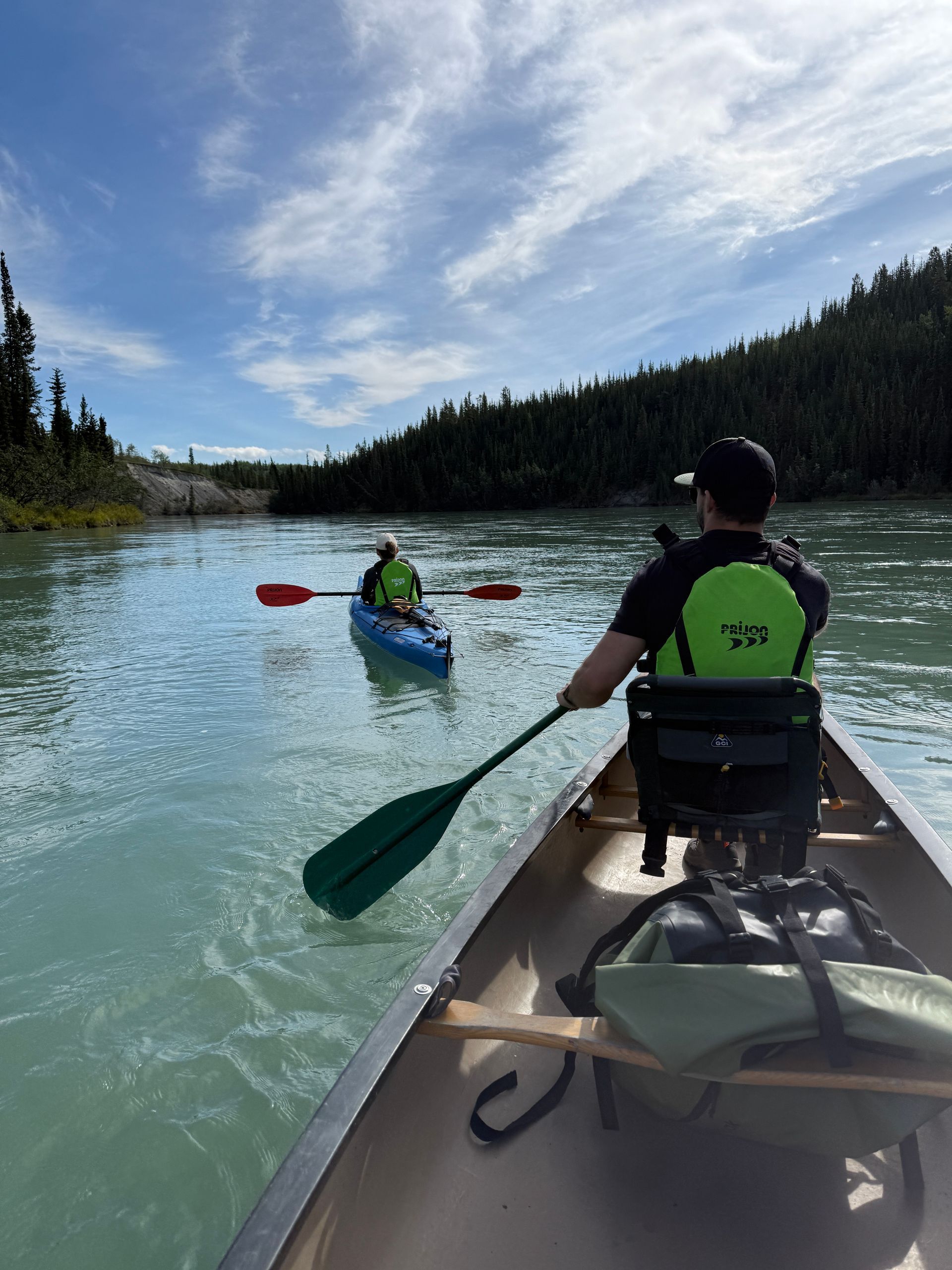 Two kayakers paddling on a calm river with forested banks under a bright blue sky