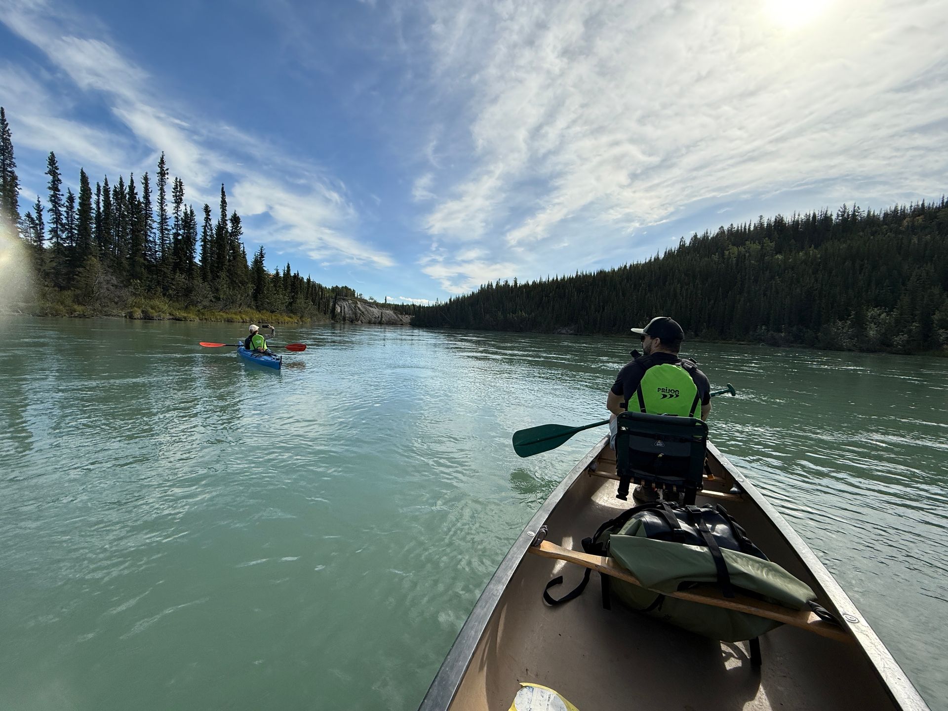 Canoer in green jacket paddles on a calm river with other kayaks, pine forest and cloudy sky ahead