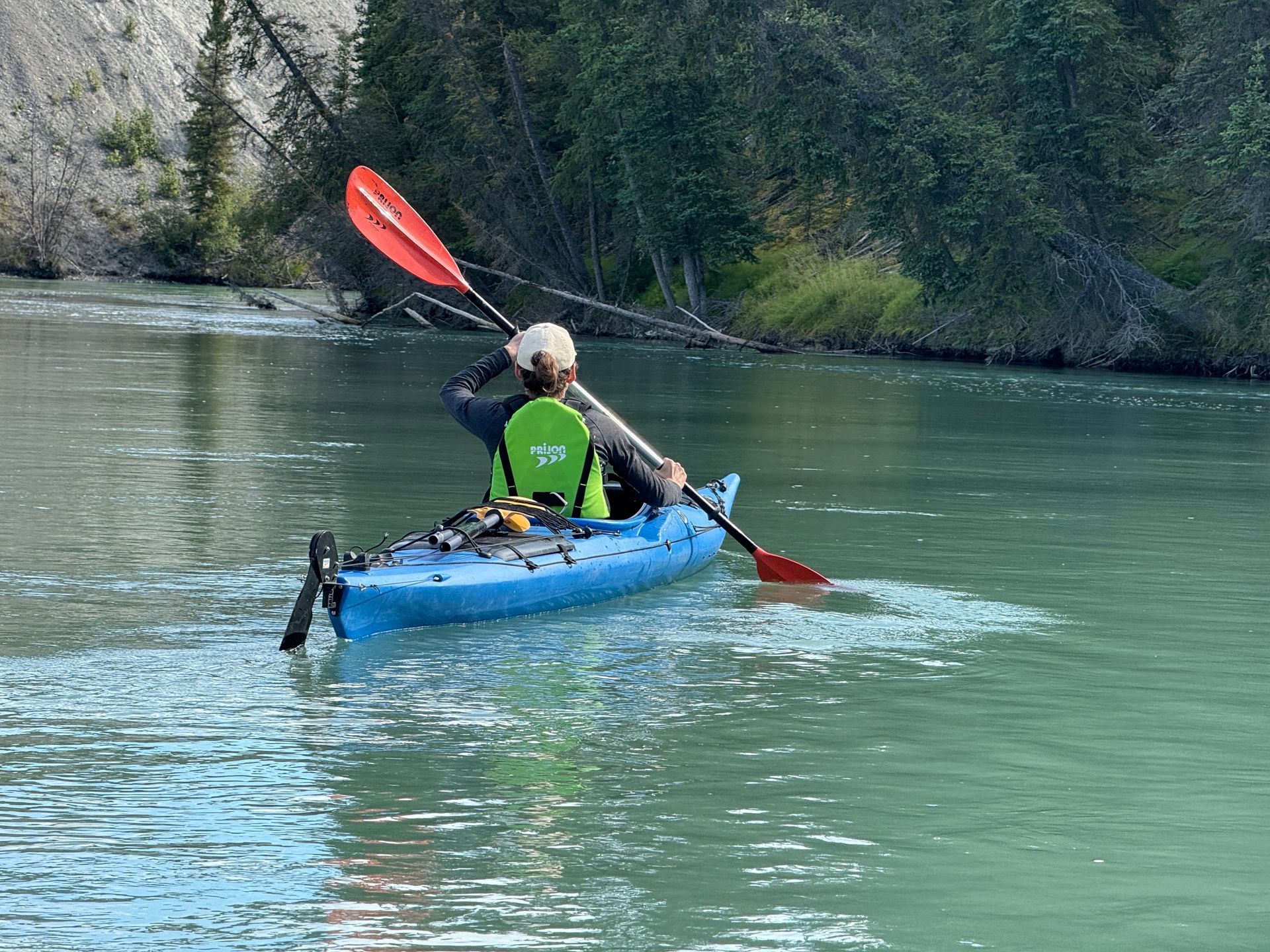 Two people kayaking on a calm green river, one paddling a blue kayak with a red paddle.