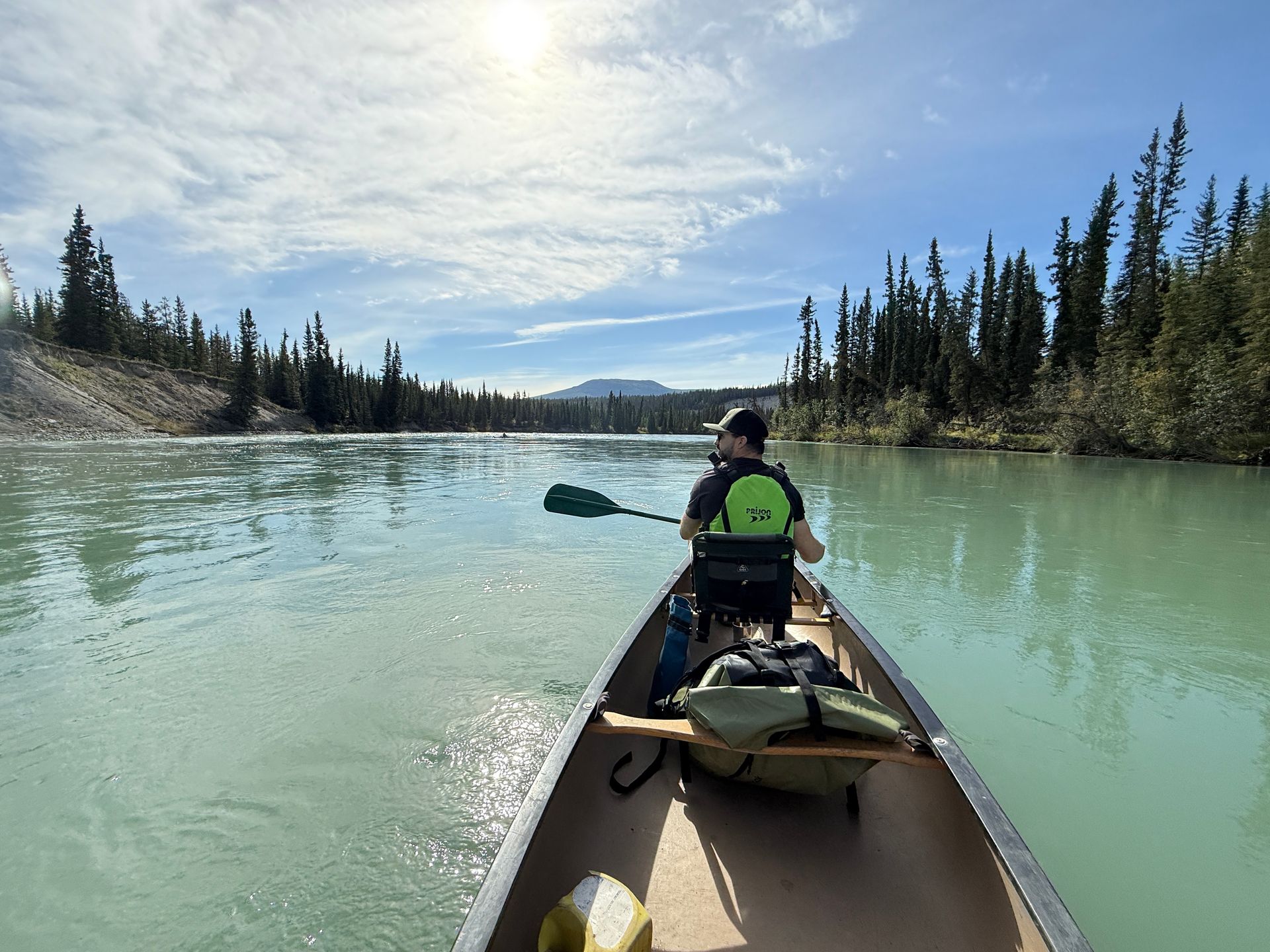 Person canoeing on a pale green river under a cloudy sky, surrounded by pine-covered banks.
