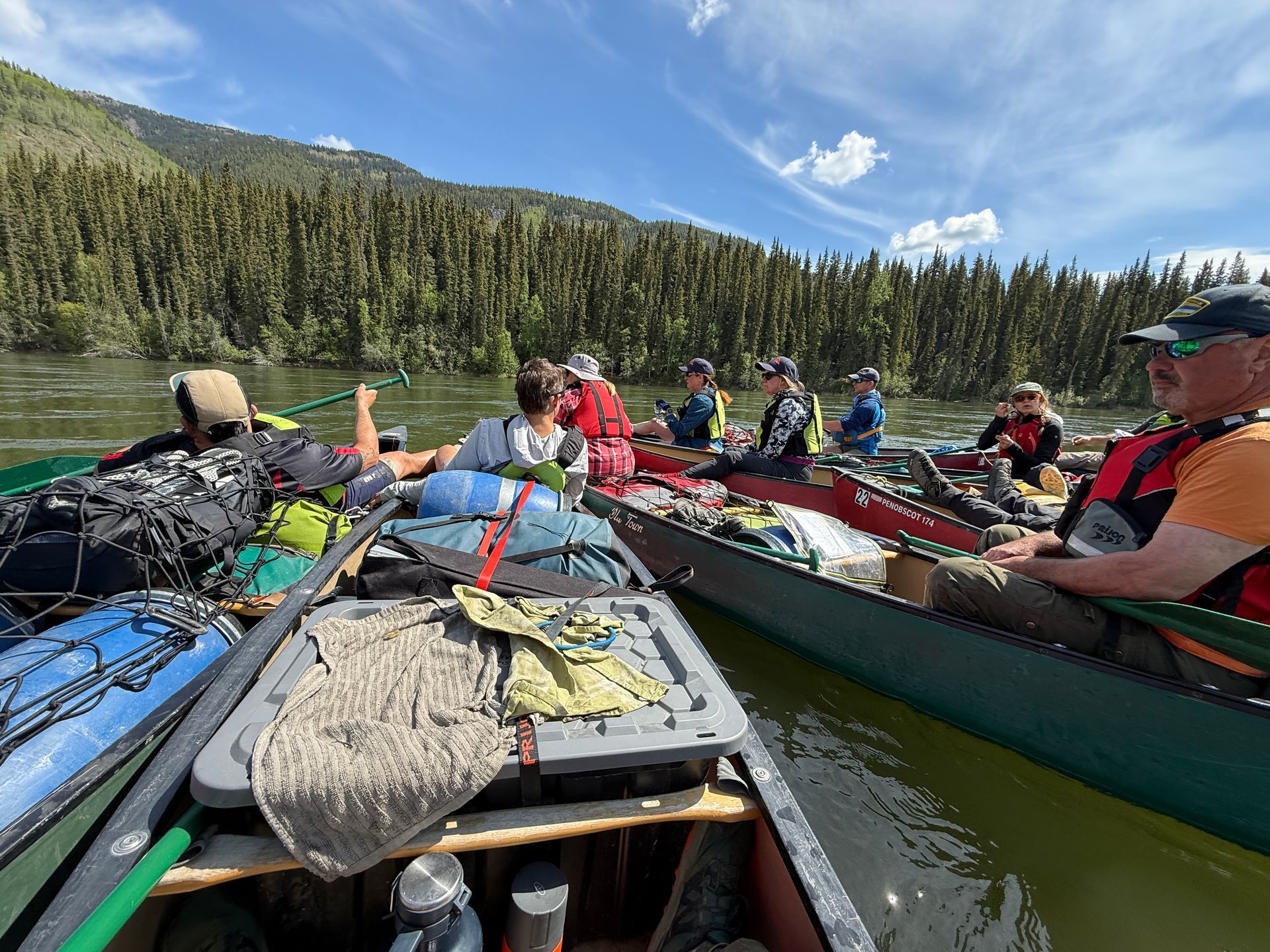 People paddling loaded canoes on a mountain lake with forested shores under a blue sky