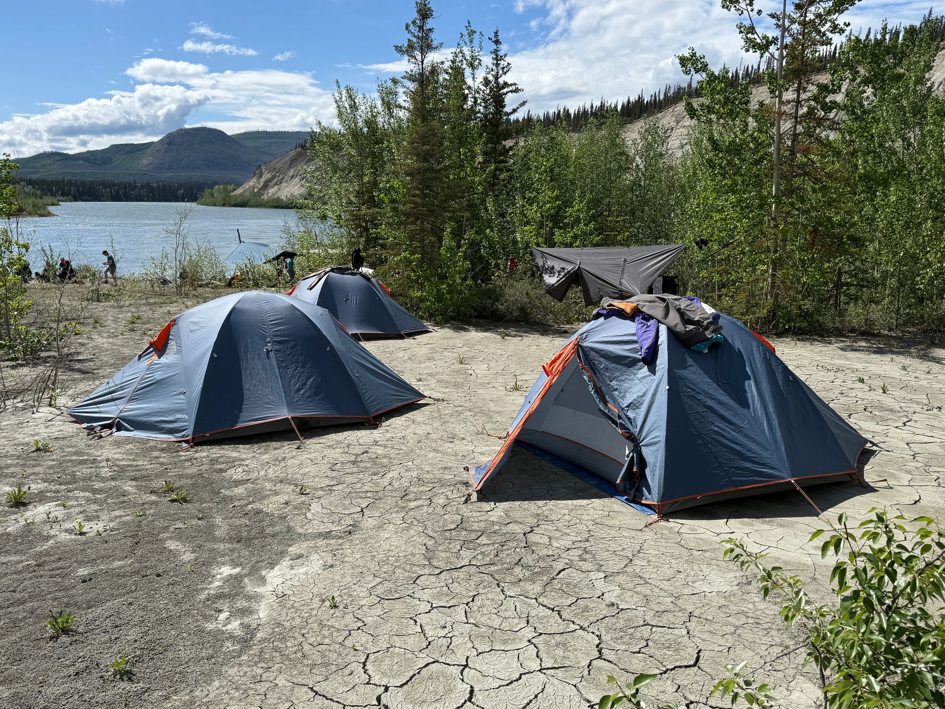 Two gray tents on cracked ground by a lake, with trees and mountains under a blue sky