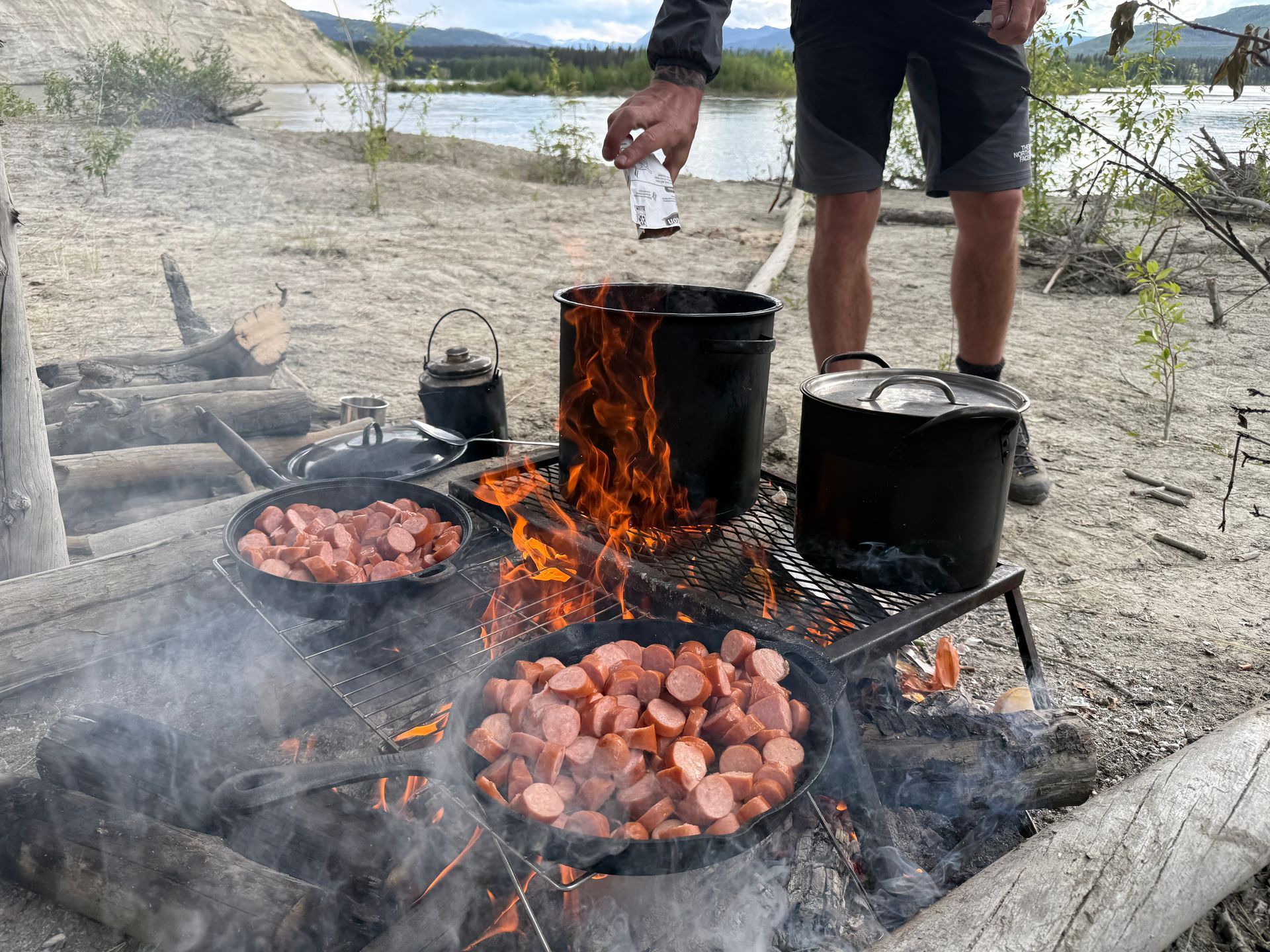 Outdoor campfire cooking with pots and pans over flames on a sandy lakeside shore