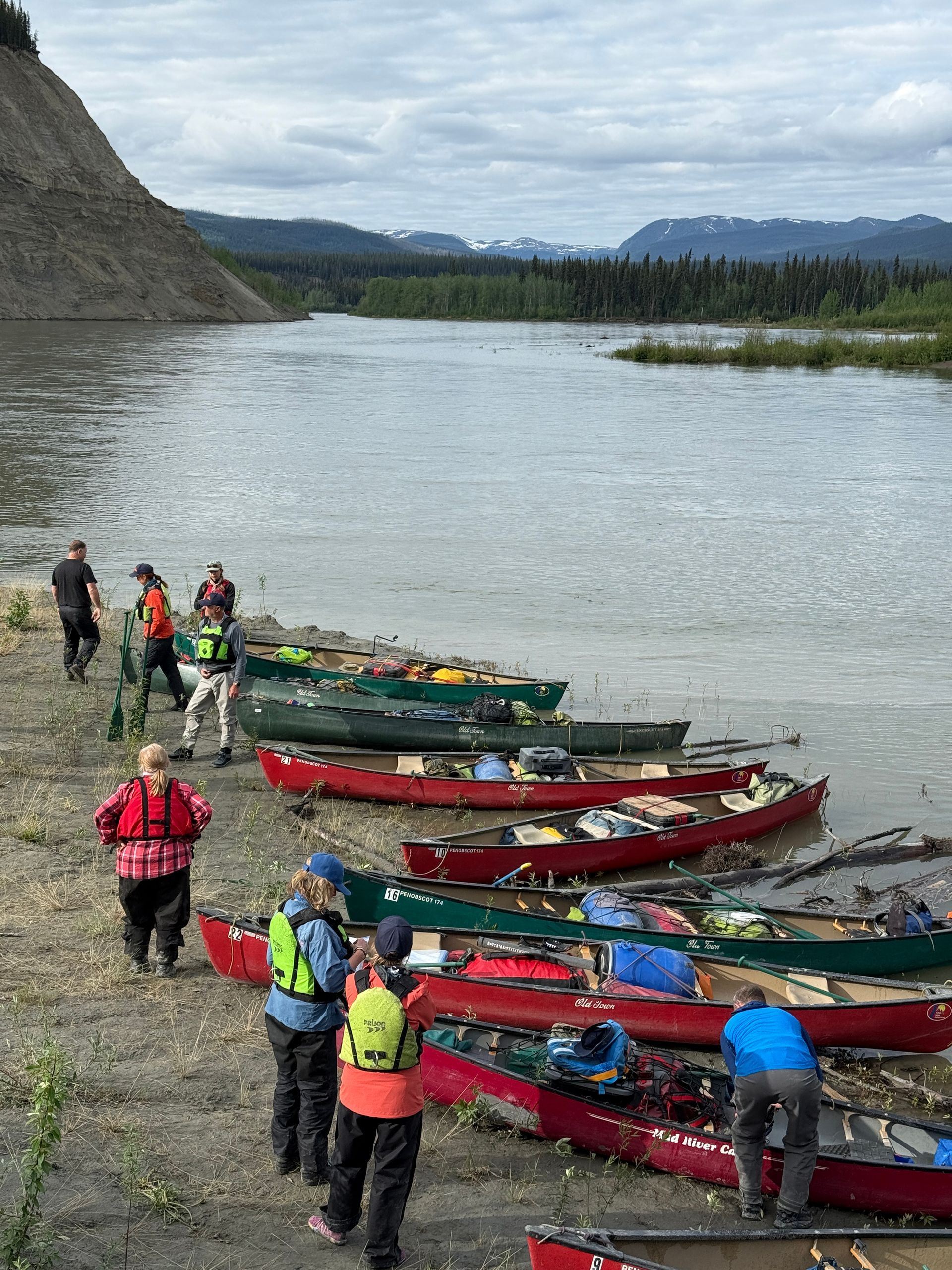 People standing by colorful canoes on a rocky riverbank beside a wide river and mountains
