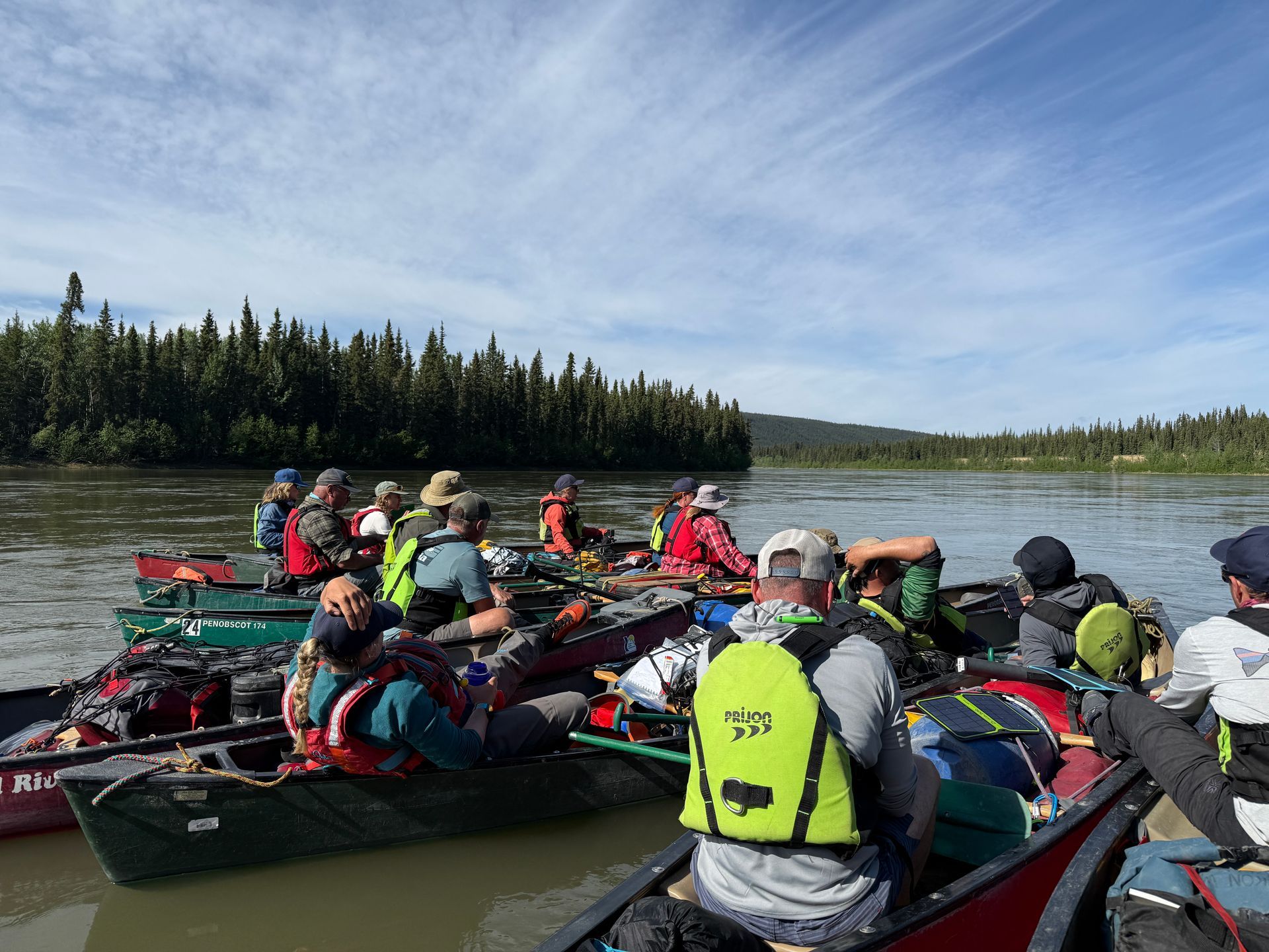 Group of people in canoes on a calm river, wearing life jackets, with forested shoreline and blue sky.