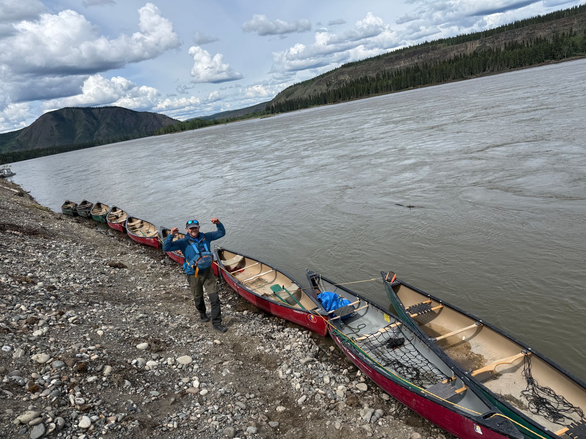 Canoes lined along a rocky riverbank, with people loading gear beside a wide river under cloudy skies