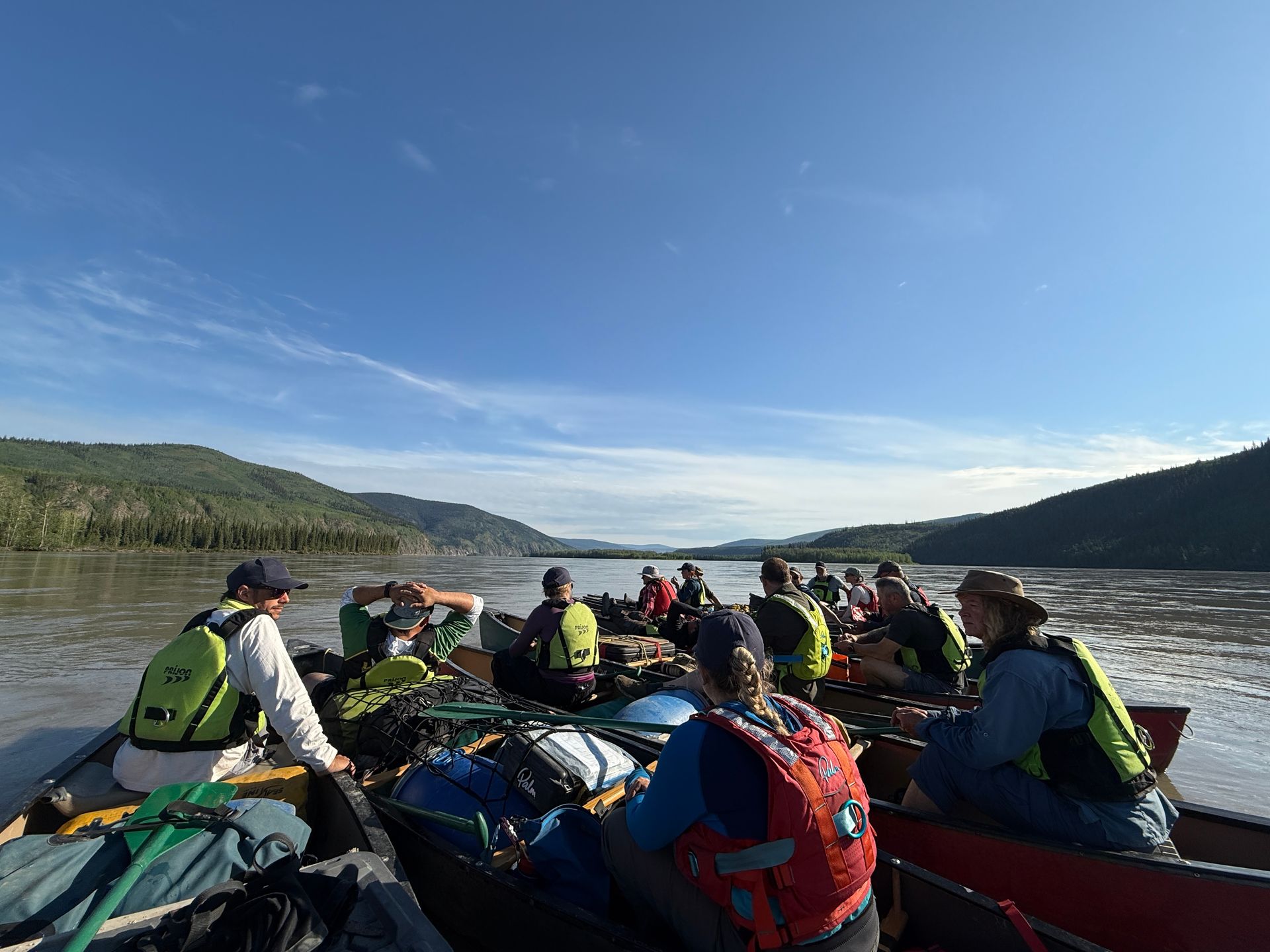 Kayakers in colorful life jackets paddle on a calm lake with forested hills and a blue sky.