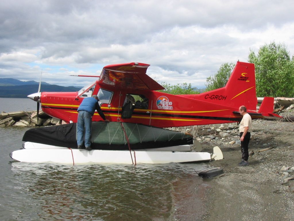 Red floatplane on a rocky shoreline with two people loading it, mountains and water in the background