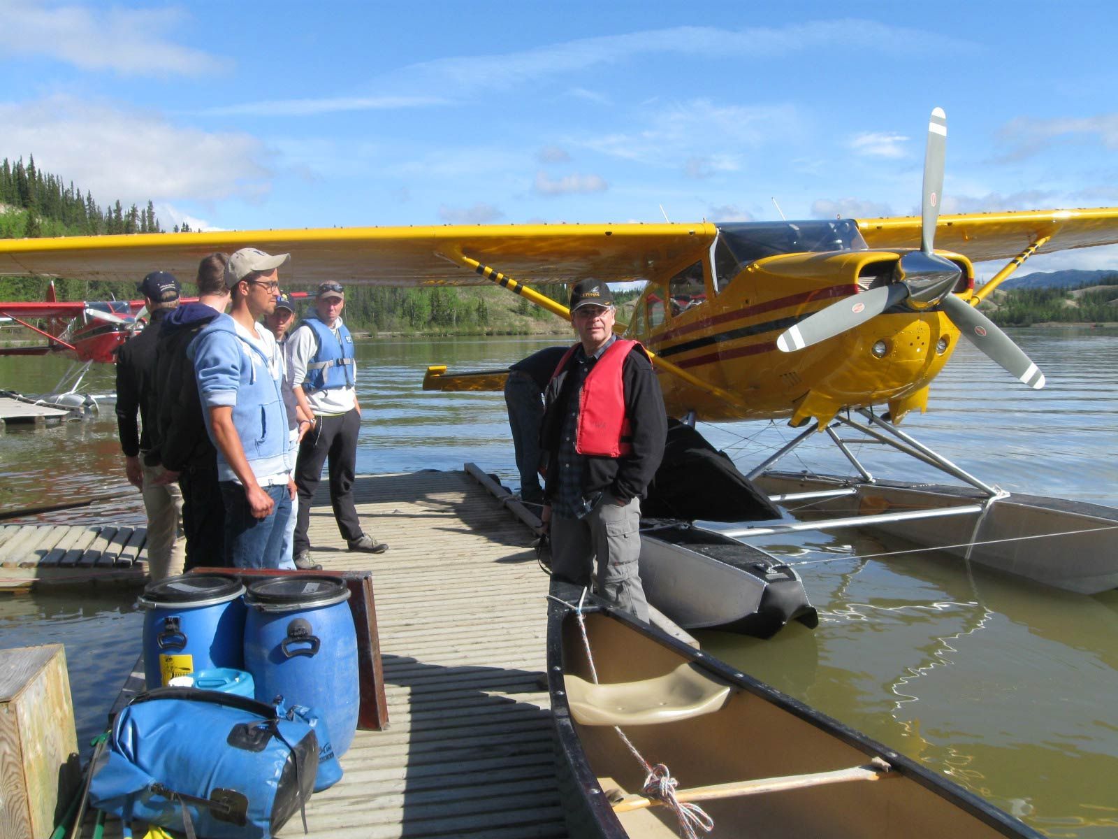 People standing beside yellow floatplane on a lakeshore with camping gear and gear bags