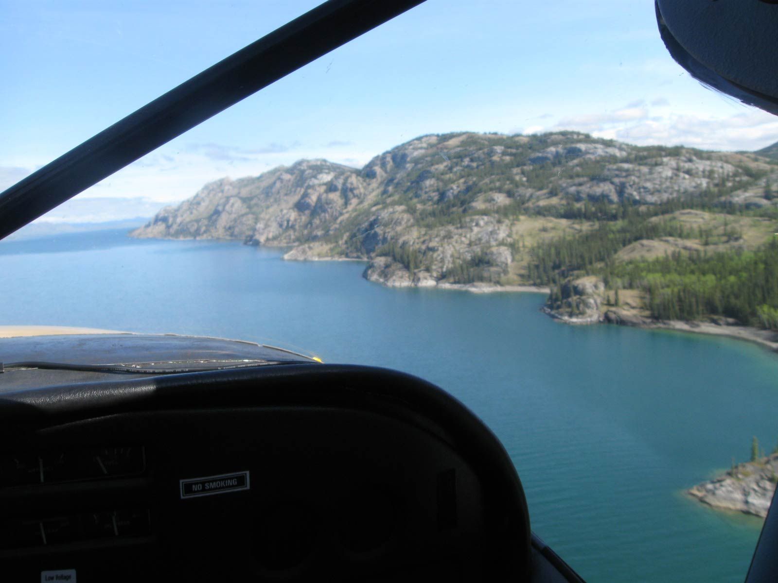 View from a cockpit over blue water toward a rocky green coastline under a clear sky