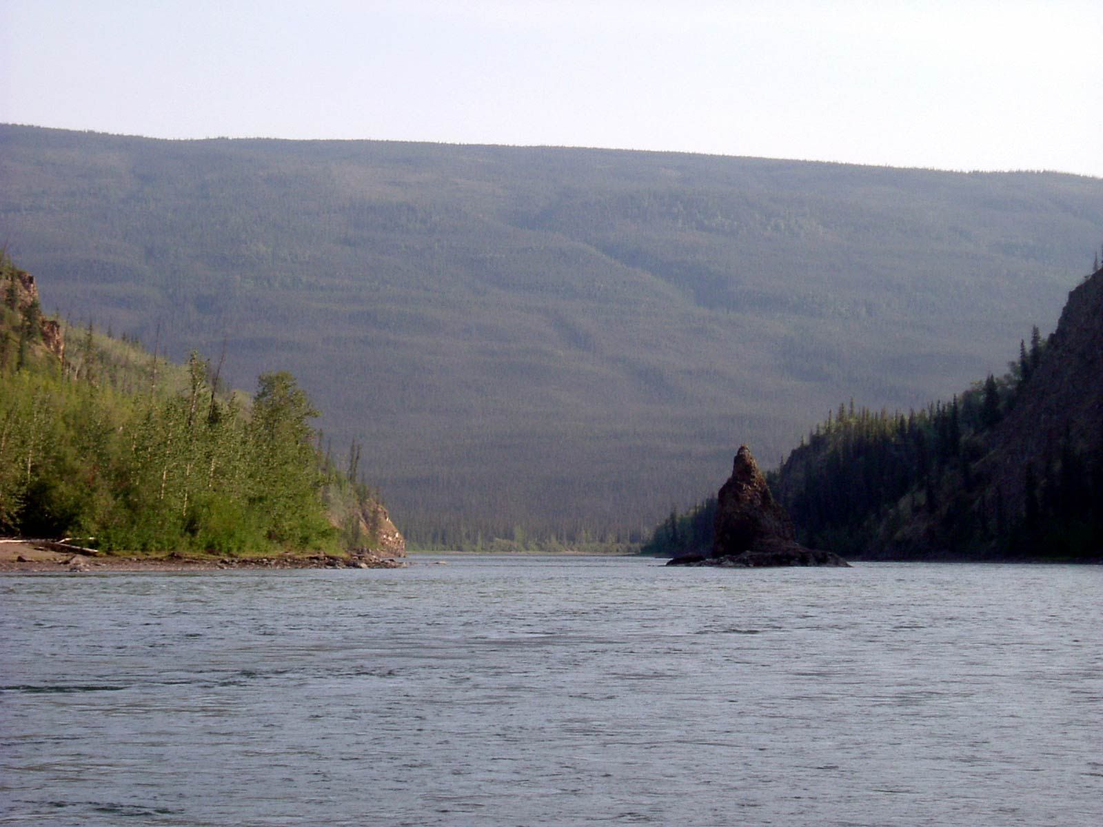 Wide river between green and dark forested hills under a hazy sky