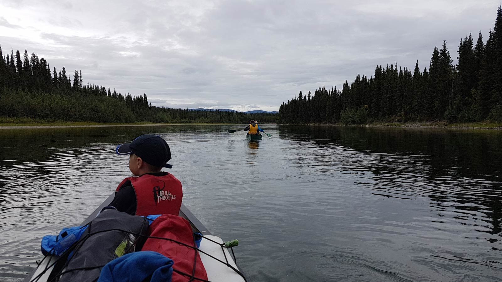 Person canoeing on a calm river with forested banks under a cloudy sky