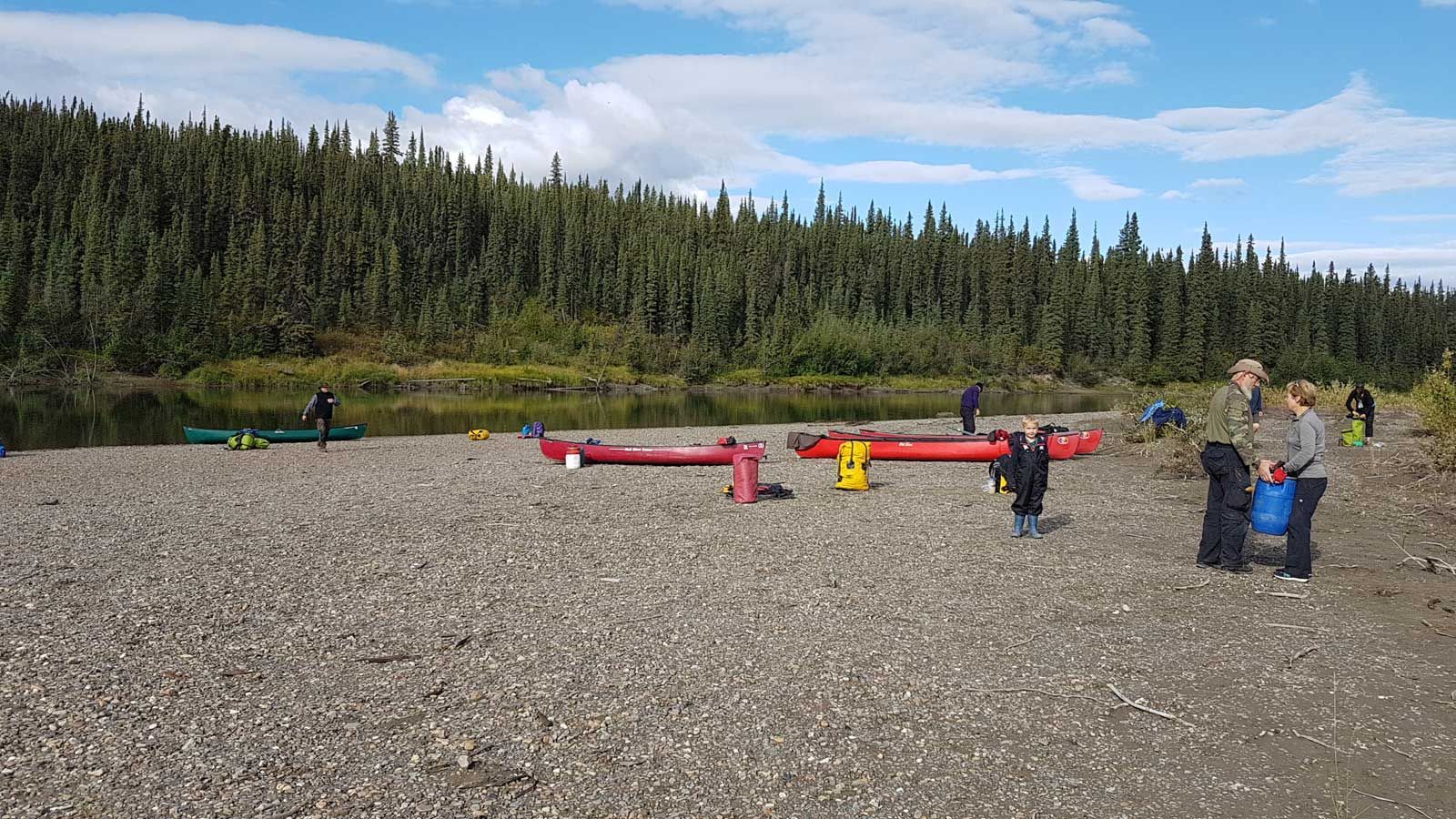 People standing by red canoes on a rocky riverbank with forest and mountains in the background