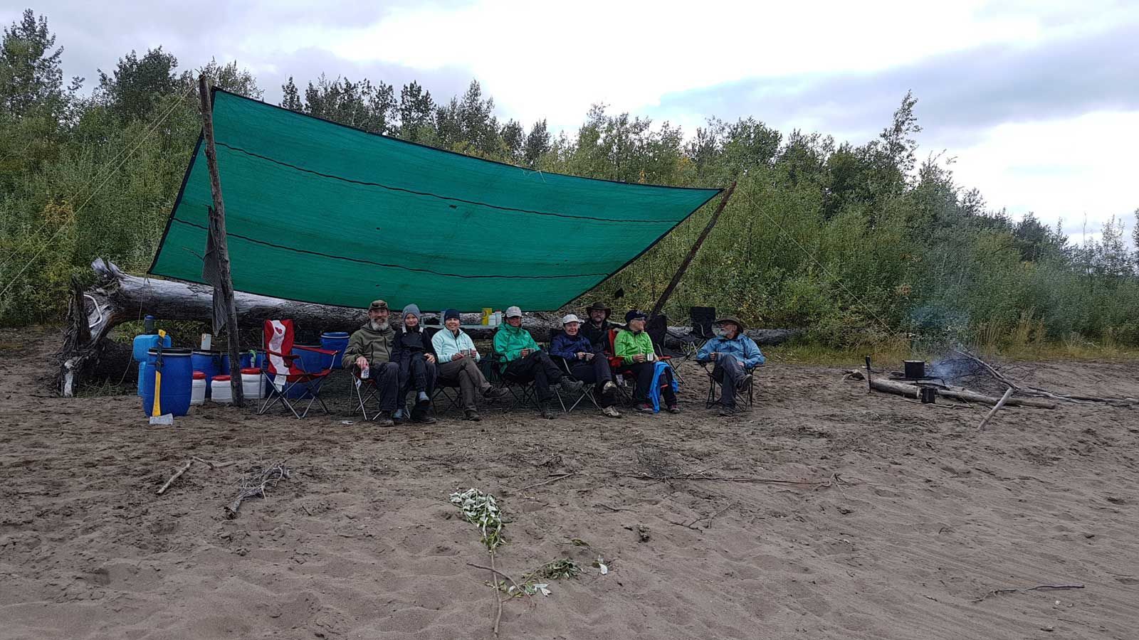 Group under a green tarp shelter in a muddy outdoor camp surrounded by trees and gear