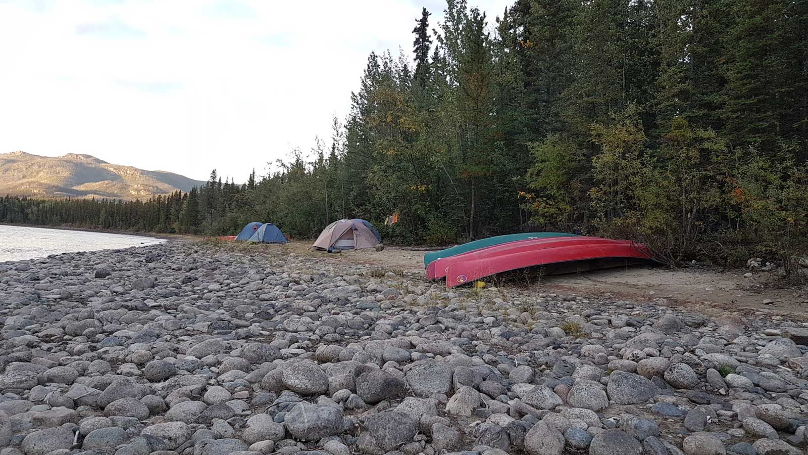 Campsite on a rocky lakeshore with two tents and a red canoe beside trees at sunset