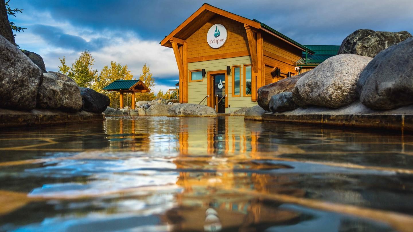 Rustic wooden cabin beside a reflective lake, with rocks, mountains, and warm sunrise light