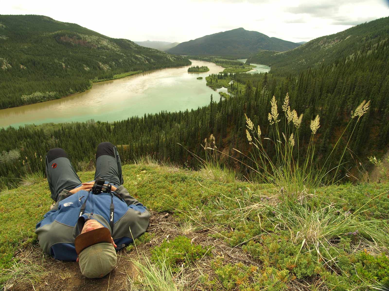 Person lying on grass overlooking a winding river valley and forested hills