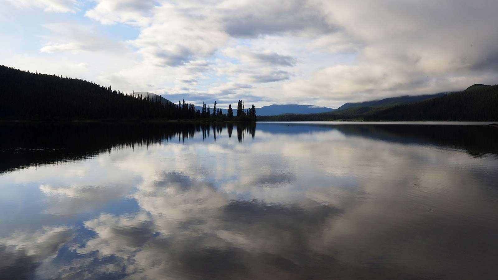 Calm lake reflecting cloudy sky and dark forested hills at sunrise or sunset