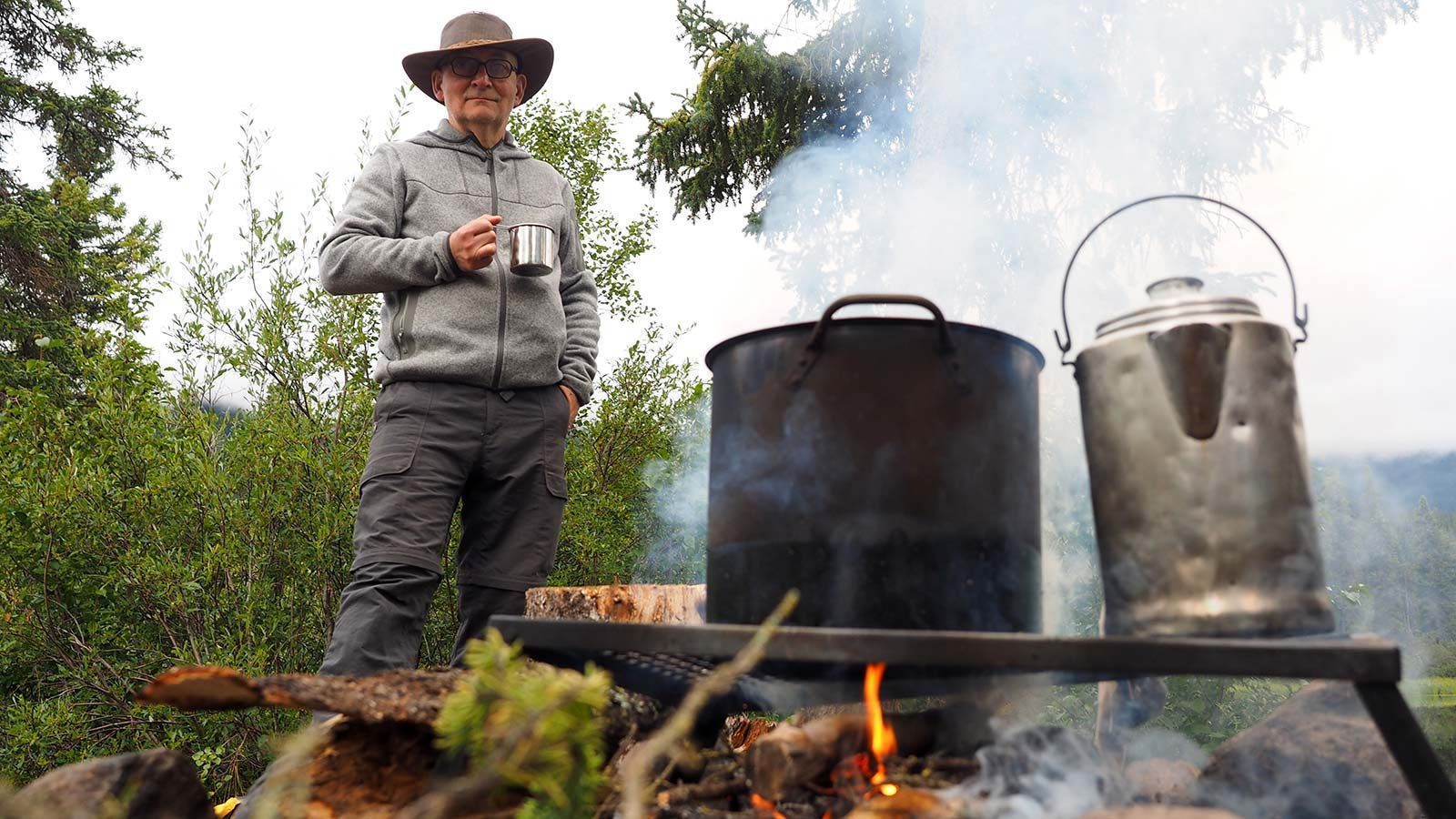 Man in a hat stands by steaming pots over an outdoor fire in a forest setting