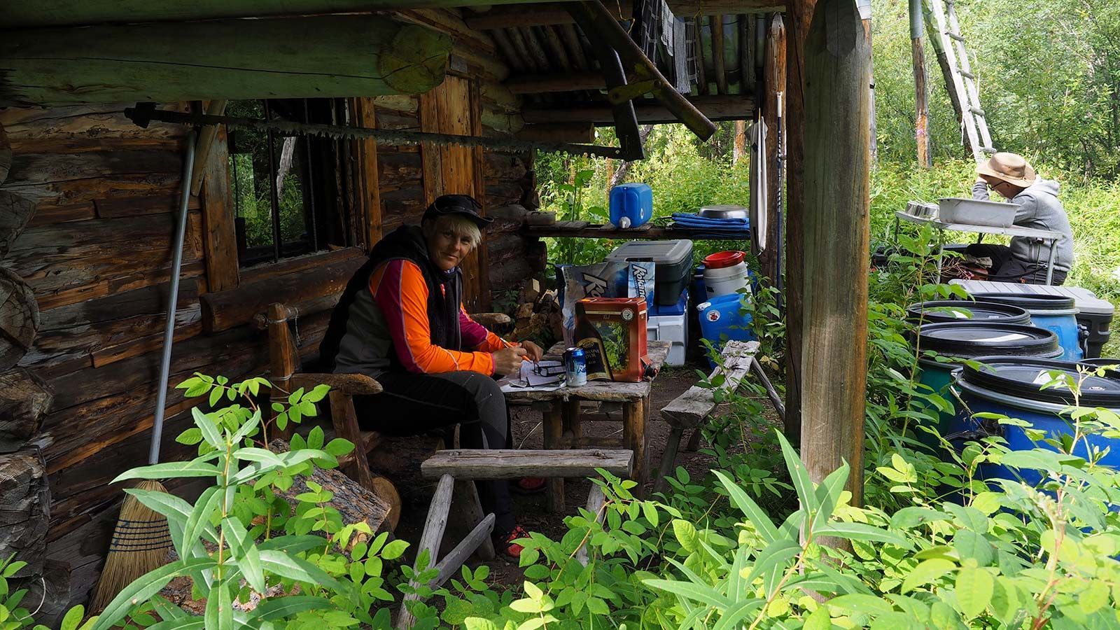 Person sitting under a rustic shelter beside stacked tires and supplies in lush greenery