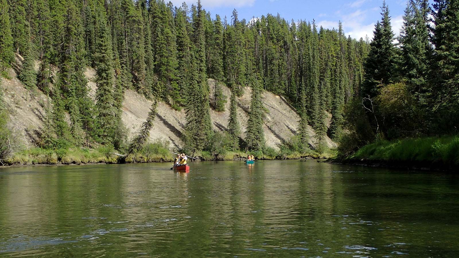 Two kayaks on a calm river beside a forested cliff under a clear blue sky